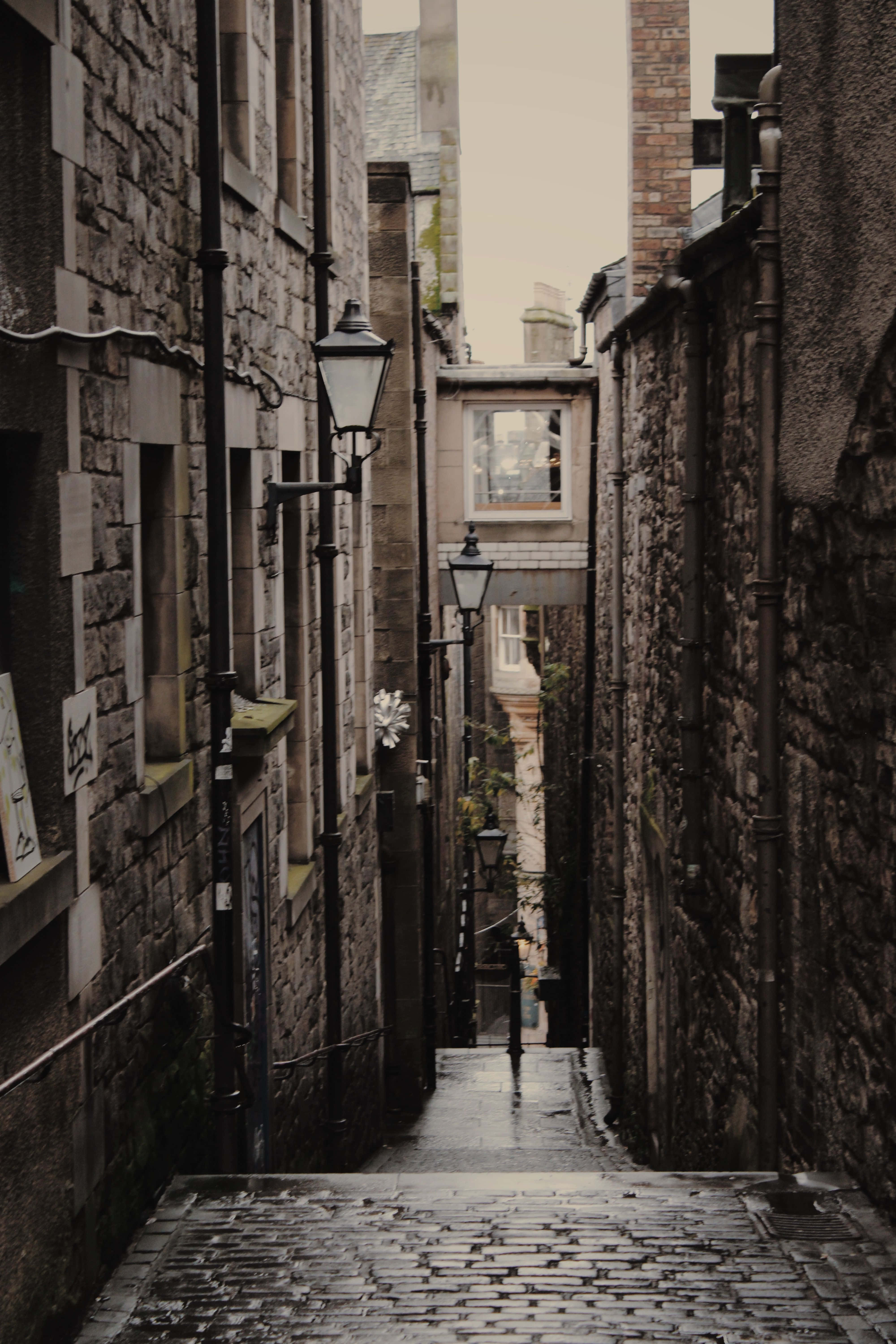 Alleyway 桌布 壁紙 — 一條狹窄的鵝卵石小巷，位於古老的石建築之間，天空多雲，配有復古路燈。