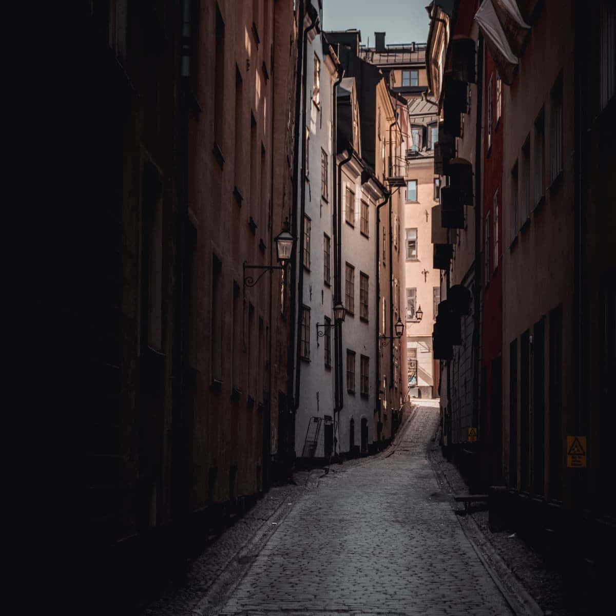 Dark Alleyway 桌布 壁紙 — 一條狹窄的鵝卵石小巷，兩旁是高聳的歷史建築，在黎明時分沐浴在柔和的金色光芒中。