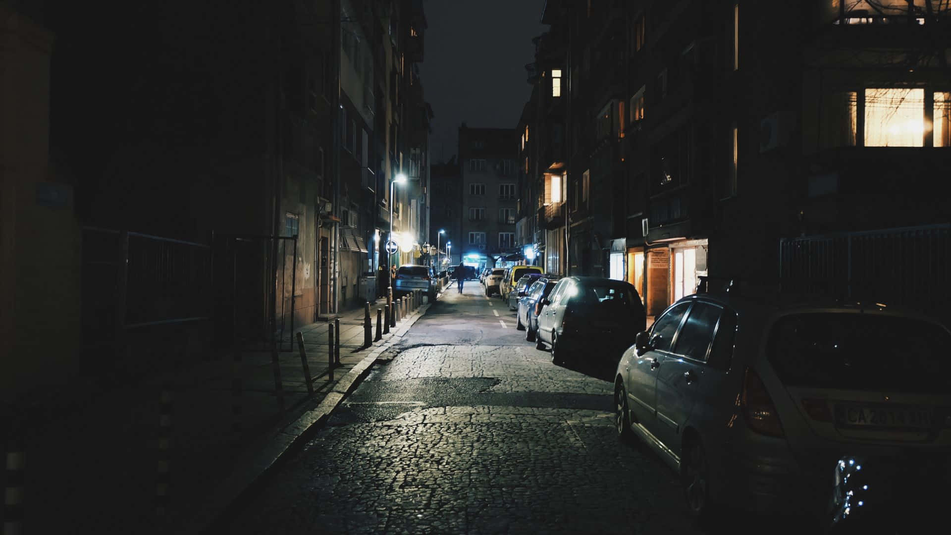 Dark Night 桌布 壁紙 — 一條狹窄的鵝卵石街道在夜晚被街燈和溫暖的窗戶光芒照亮，兩旁停滿了汽車。