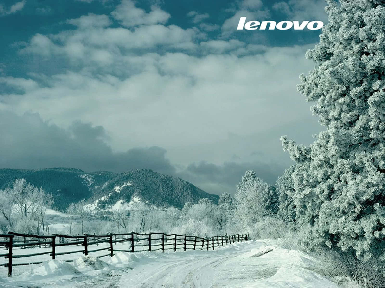 Lenovo 桌布 壁紙 — 一條白雪覆蓋的鄉村道路蜿蜒經過結霜的樹木和木柵欄，通往遠處白雪覆蓋的山脈，天空多雲的冬日天空。
