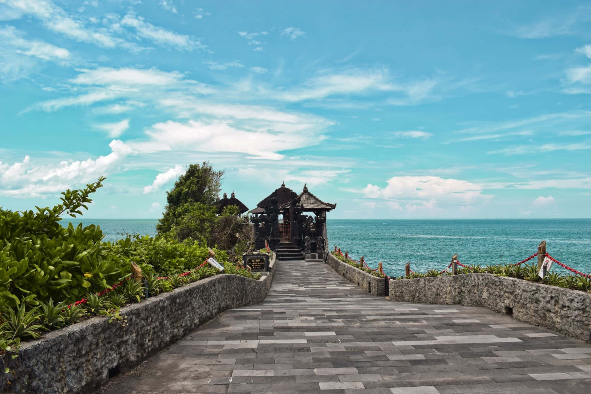 Bali 桌布 壁紙 — 一條石徑通往一座傳統峇里寺廟，寺廟坐落在懸崖上，俯瞰著湛藍的天空下碧綠色的海洋。
