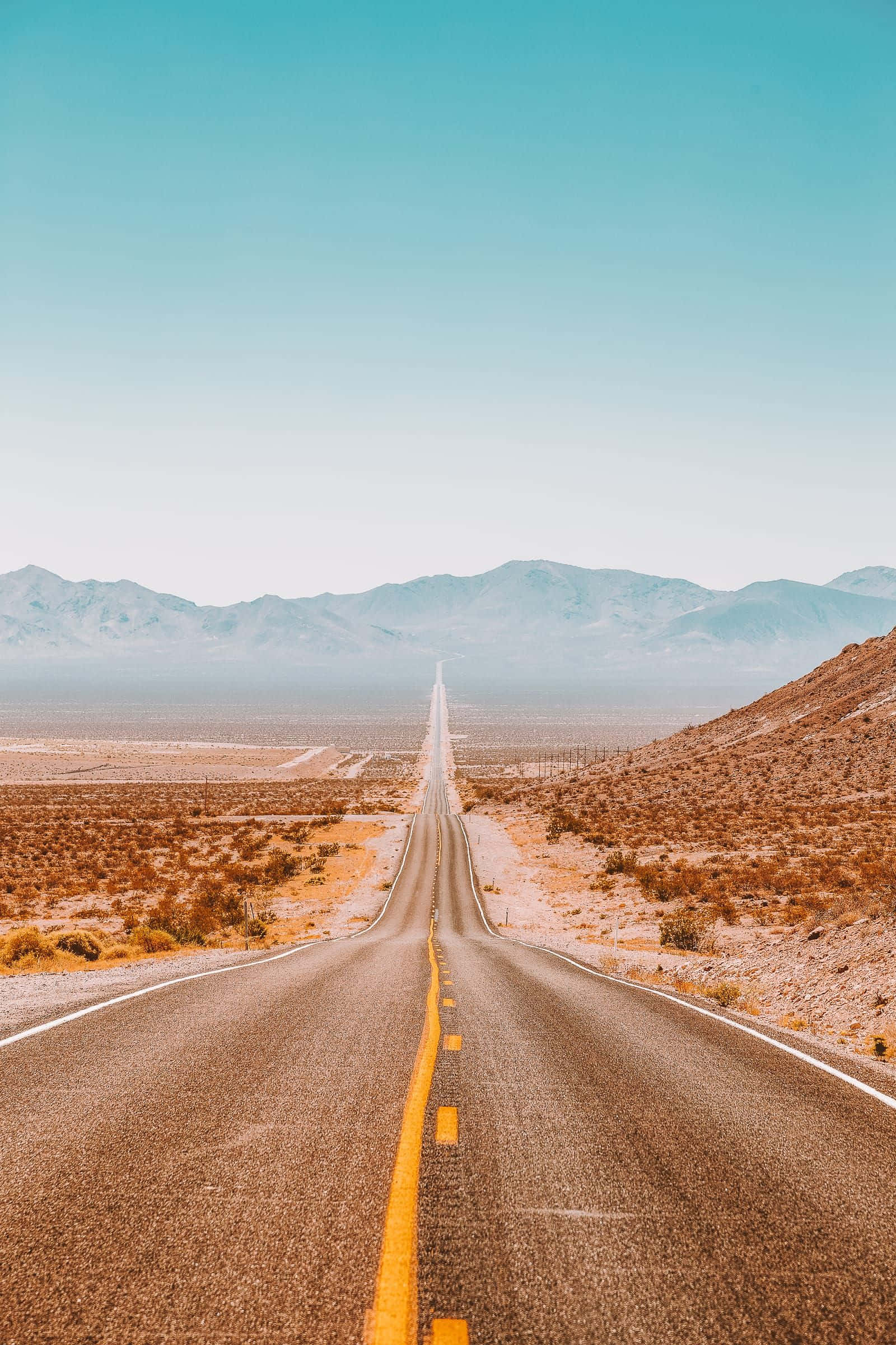 Travel Aesthetic Phone Empty Road 壁紙 — 一條筆直的沙漠高速公路在清澈的藍天下延伸至遠山。