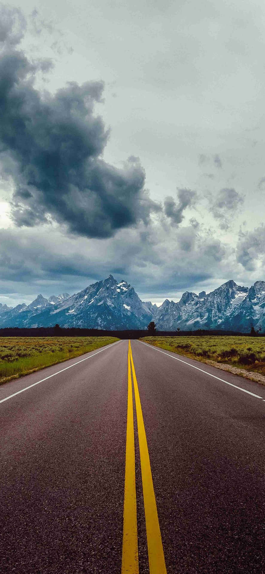 iPhone 13 Mini 桌布 壁紙 — 一條筆直的瀝青道路，帶有黃色線條，在戲劇性的多雲天空下延伸向積雪覆蓋的山脈。