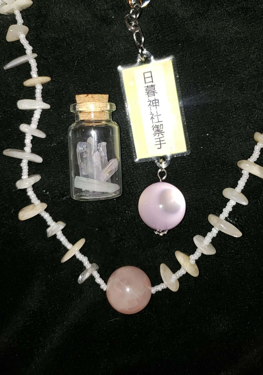  壁紙 — 一條精緻的項鍊，帶有白色珠子和粉紅色寶石，旁邊放著一個小水晶罐和一個寫著「日暮神社御手」的標籤，背景是黑色天鵝絨。