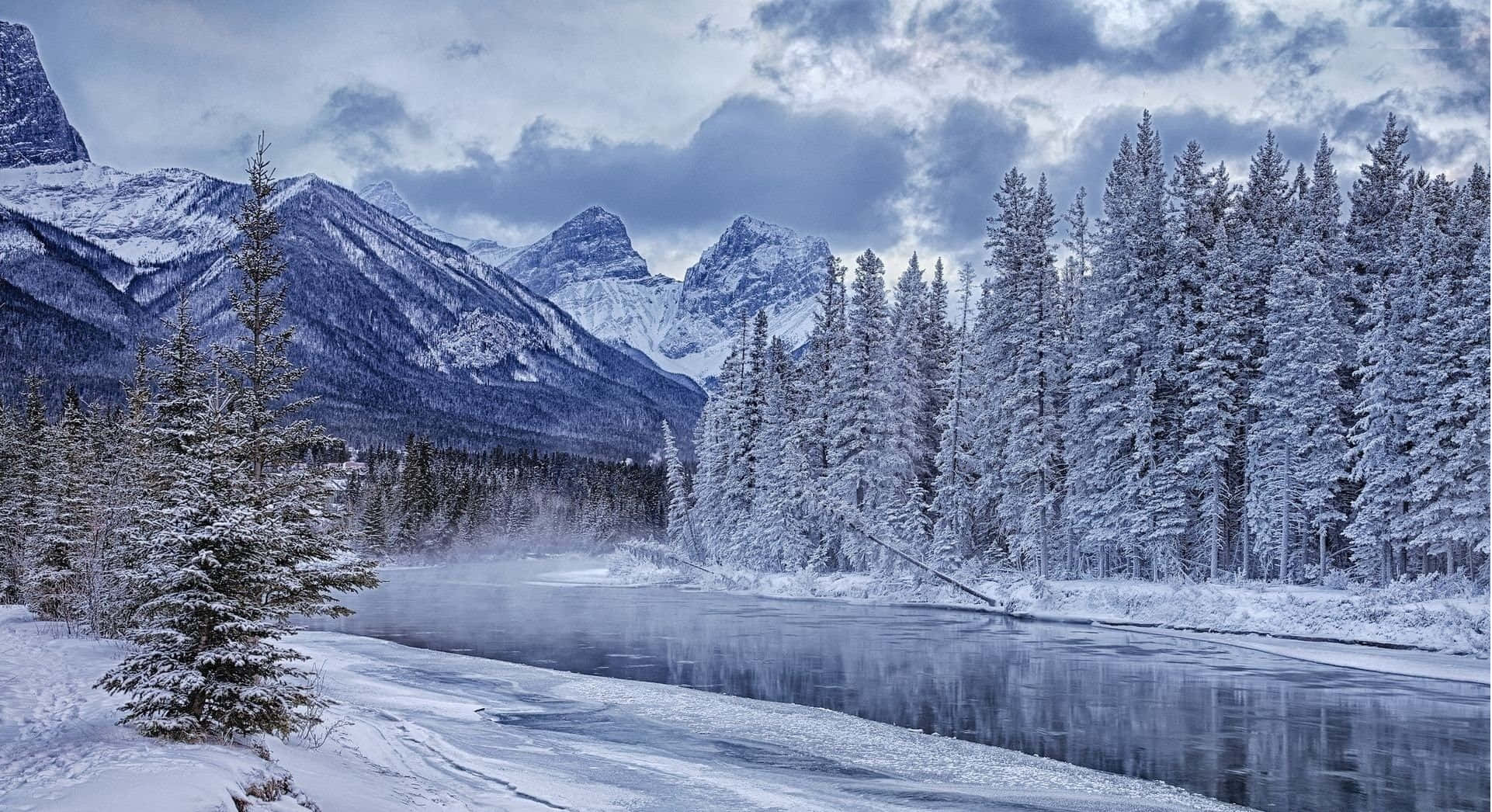 Snowy Landscape 桌布 壁紙 — 一條結冰的河流蜿蜒穿過白雪覆蓋的森林，雪覆蓋的山脈在陰沉的冬日天空下。