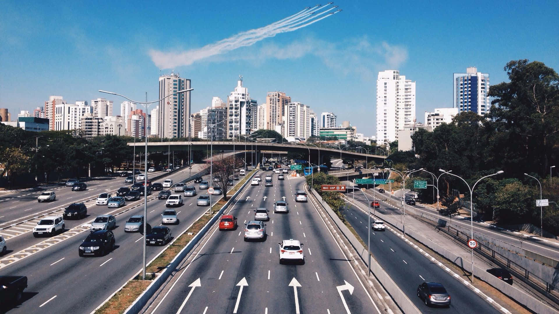 Beautiful Travel 桌布 壁紙 — 一條繁忙的多線高速公路充滿汽車，在晴朗藍天和飛機尾跡下穿過現代城市景觀。