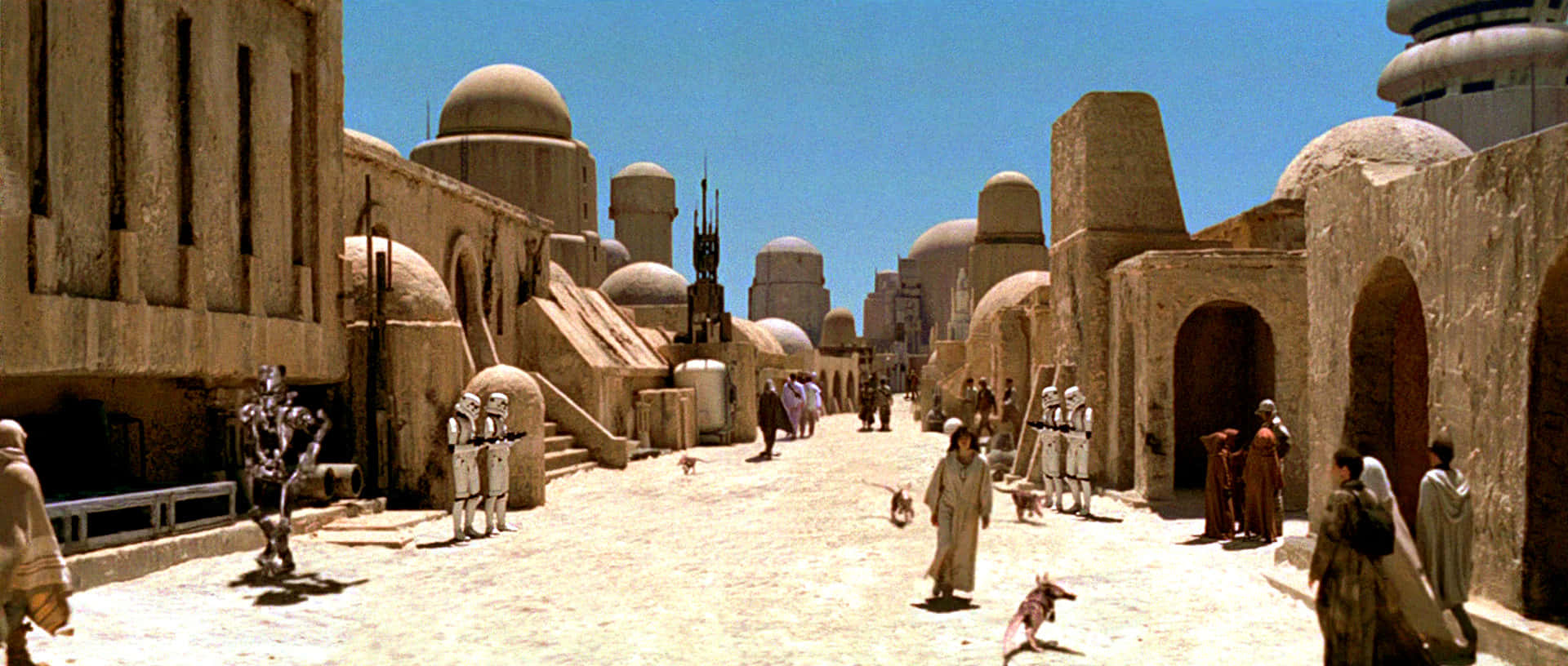 Mos Eisley 桌布 壁紙 — 一條繁忙的沙漠城市街道，有砂岩建築、圓頂和穿著長袍的人們在晴朗的藍天下行走。