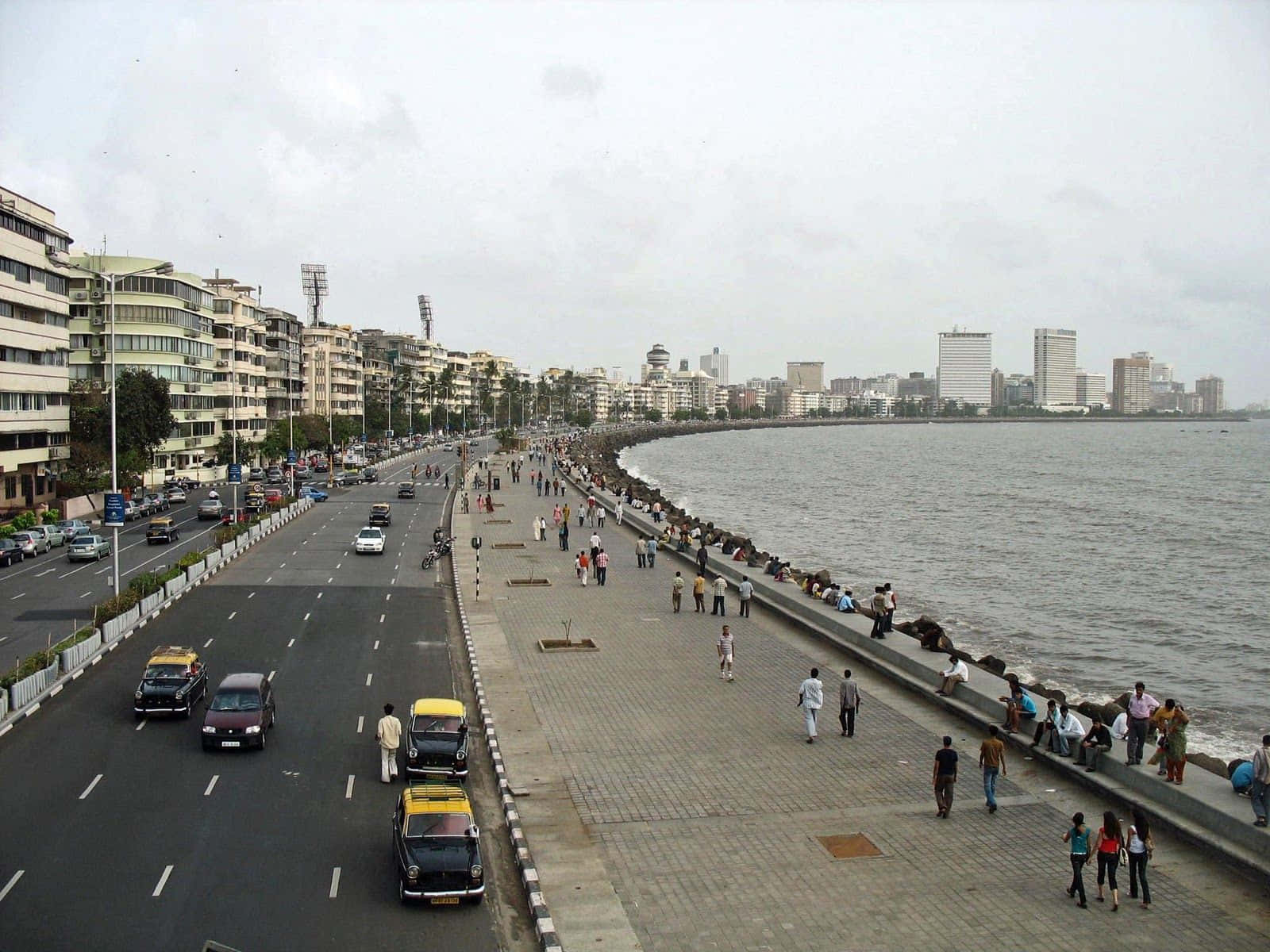 Mumbai 桌布 壁紙 — 一條繁忙的海濱長廊，行人與黃色的士沿著海邊的寬闊道路行駛，天空多雲。