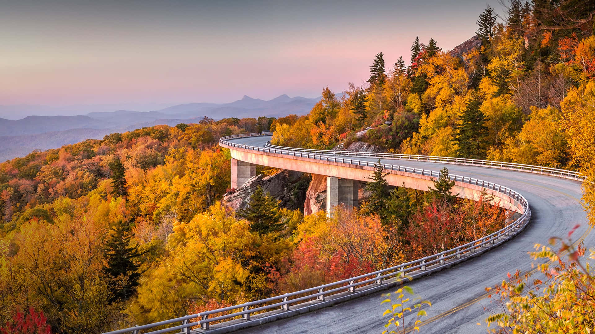 Blue Ridge Parkway 桌布 壁紙 — 一條蜿蜒的山路優雅地繞過懸崖邊，周圍是充滿活力的秋葉，在柔和的粉紅色和橙色日落天空下。