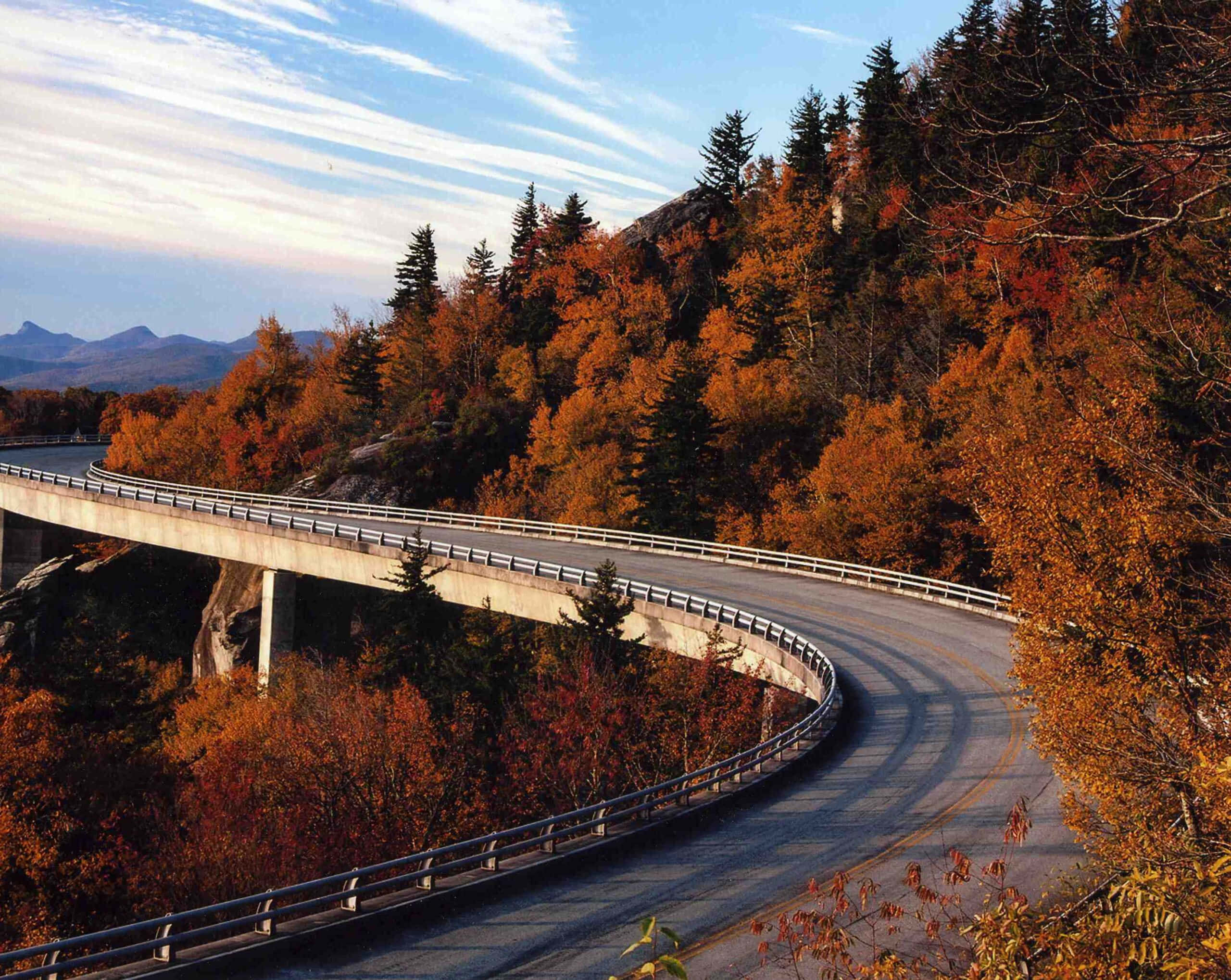 Blue Ridge Parkway 桌布 壁紙 — 一條蜿蜒的山路在柔和的藍天和薄雲下，優雅地穿過色彩鮮豔的秋葉。