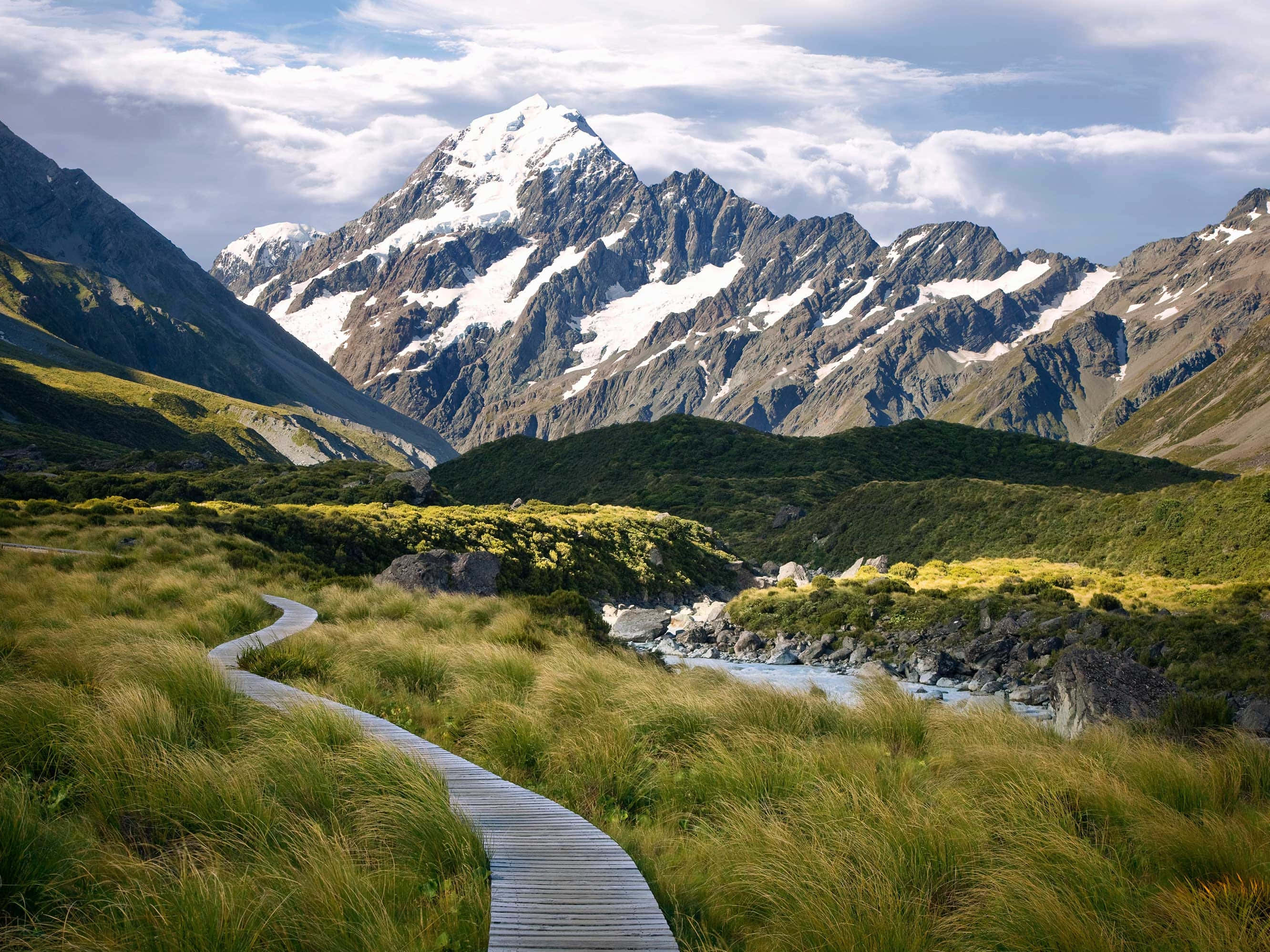 New Zealand 桌布 壁紙 — 一條蜿蜒的木棧道穿過河邊的金色草叢，通向部分多雲天空下的雪山。