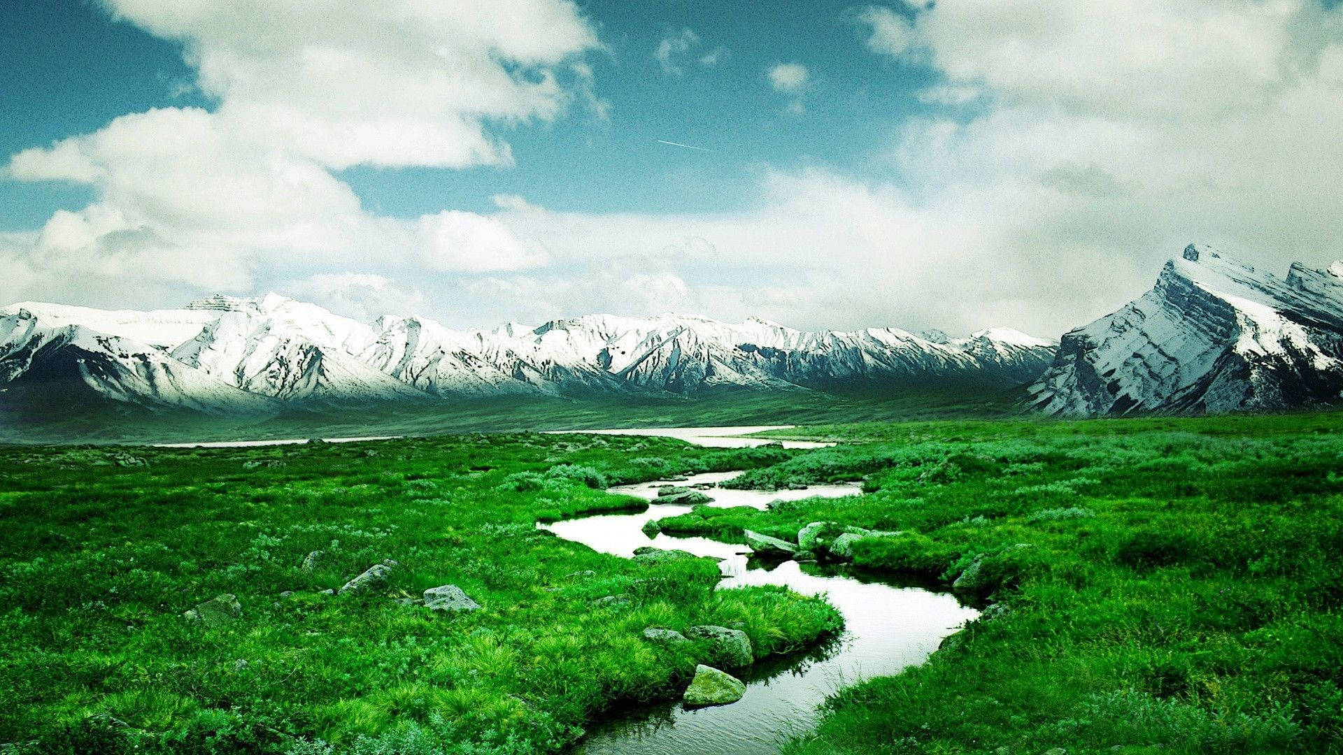 自然 桌布 壁紙 — 一條蜿蜒的河流流經充滿活力的綠色草地，白雪覆蓋的山脈在部分多雲的天空下聳立。