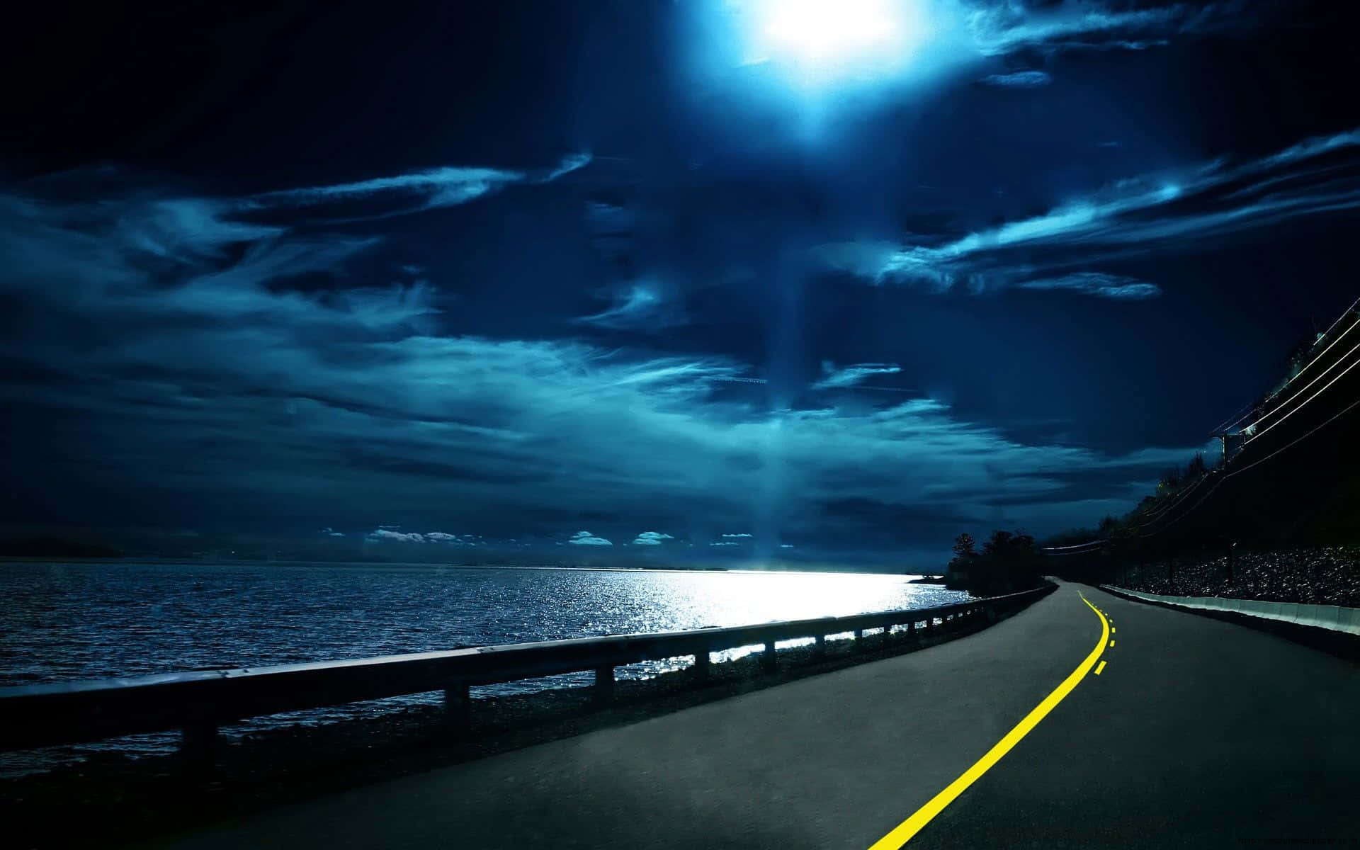 Chilly Night 桌布 壁紙 — 一條蜿蜒的海岸道路在夜晚沿著黑暗的大海彎曲，在明亮的月亮下，月亮在雲層和水面上投射出空靈的藍光。