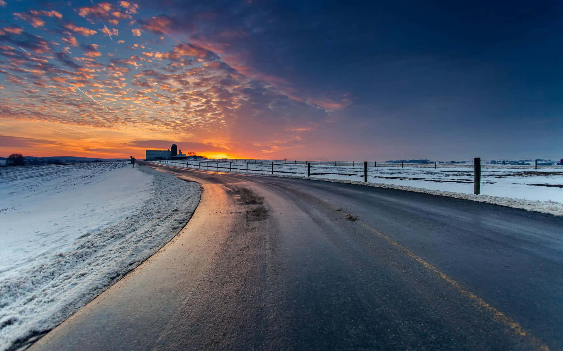 Sunset Landscape 桌布 壁紙 — 一條蜿蜒的道路在日出時穿過雪地，遠處農場上方有鮮豔的橙色和紫色雲彩。