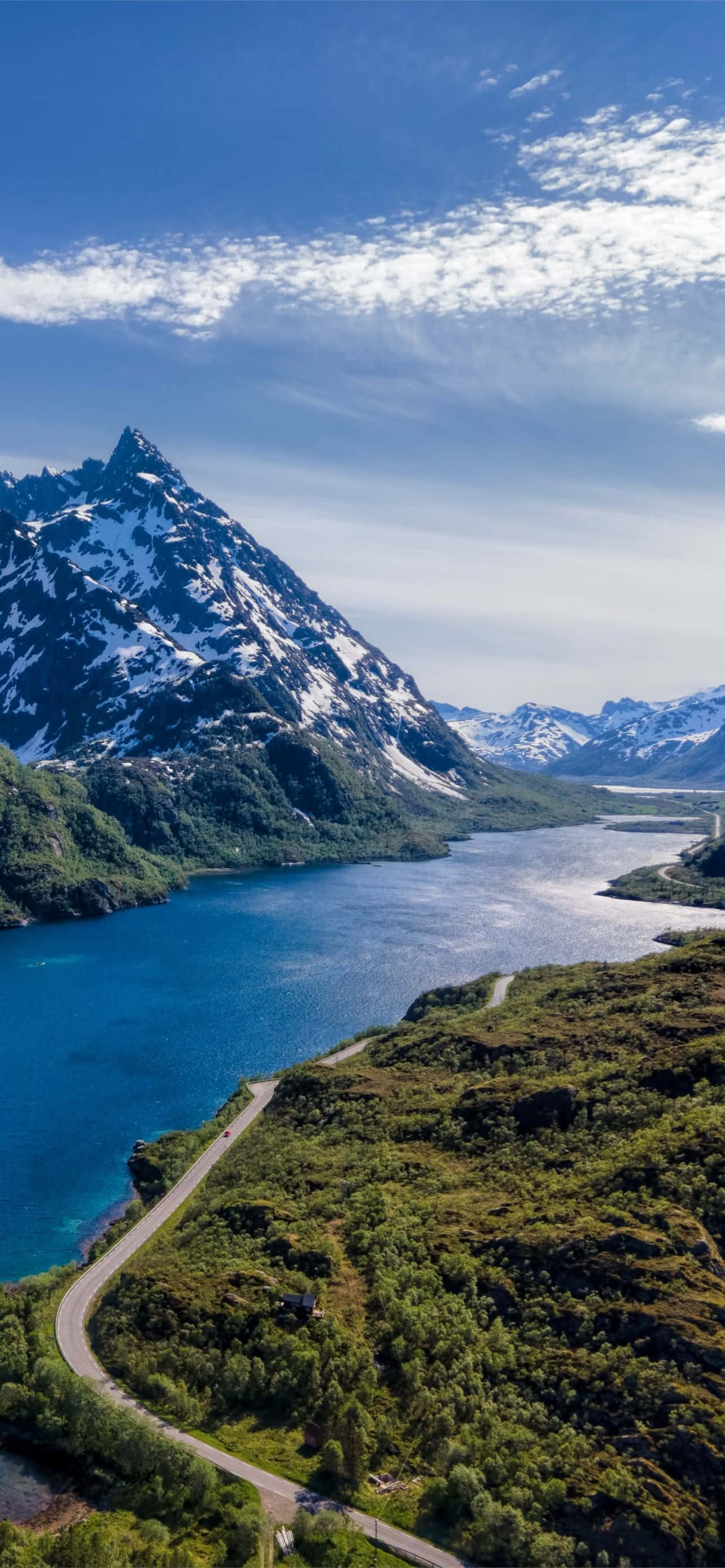 Norway 桌布 壁紙 — 一條蜿蜒的道路擁抱著充滿活力的藍色峽灣，下方是白雪覆蓋的山脈，上方是明亮的藍天和縷縷白雲。