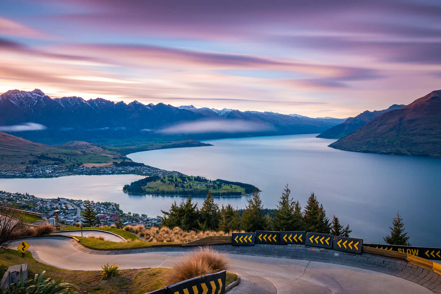 New Zealand 桌布 壁紙 — 一條蜿蜒的道路沿著山坡彎曲，俯瞰黃昏時寧靜的湖泊，遠處是白雪覆蓋的山峰。