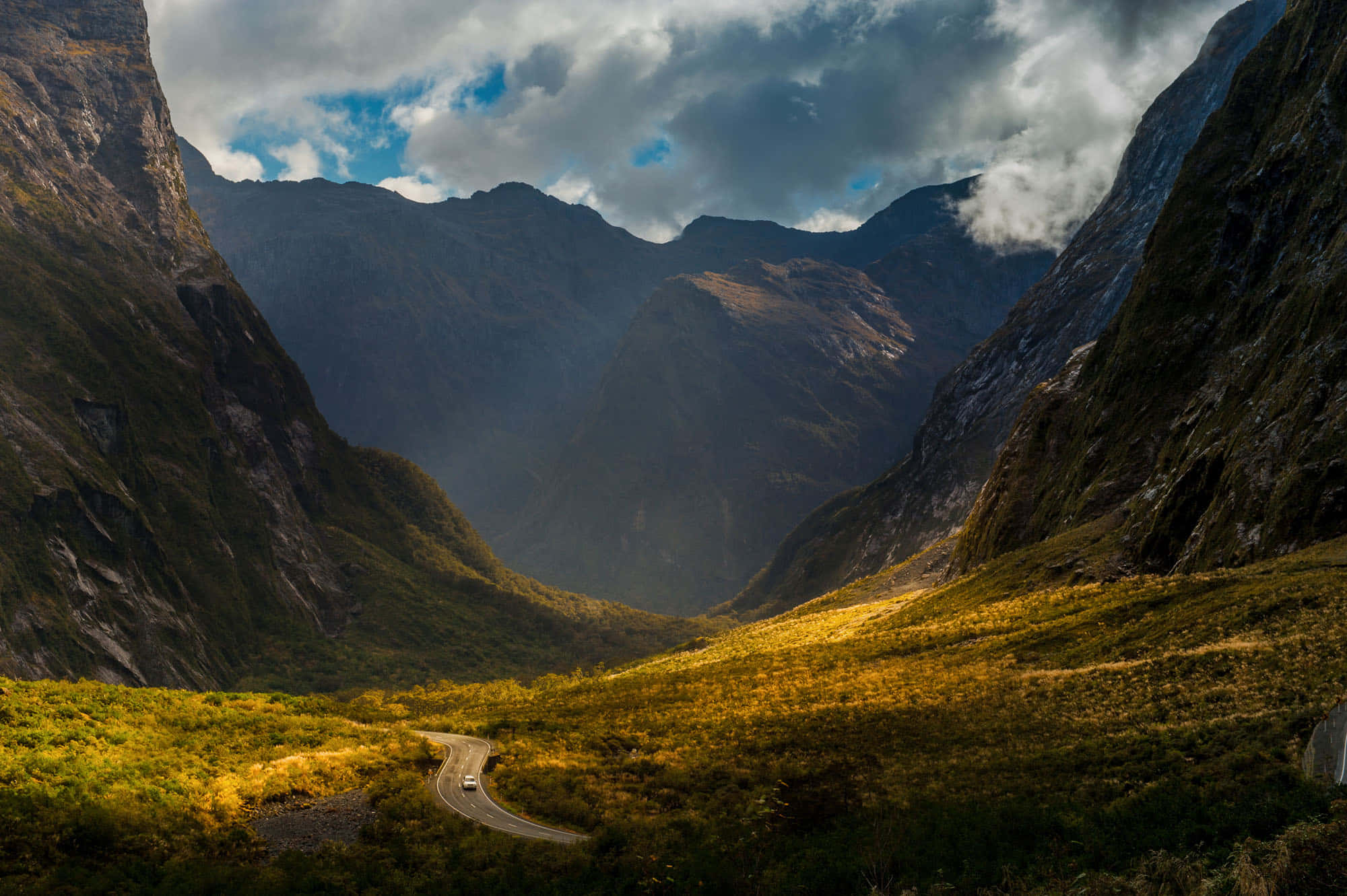 New Zealand 桌布 壁紙 — 一條蜿蜒的道路穿過一個戲劇性的山谷，沐浴在穿透暴風雲層的金色陽光中。