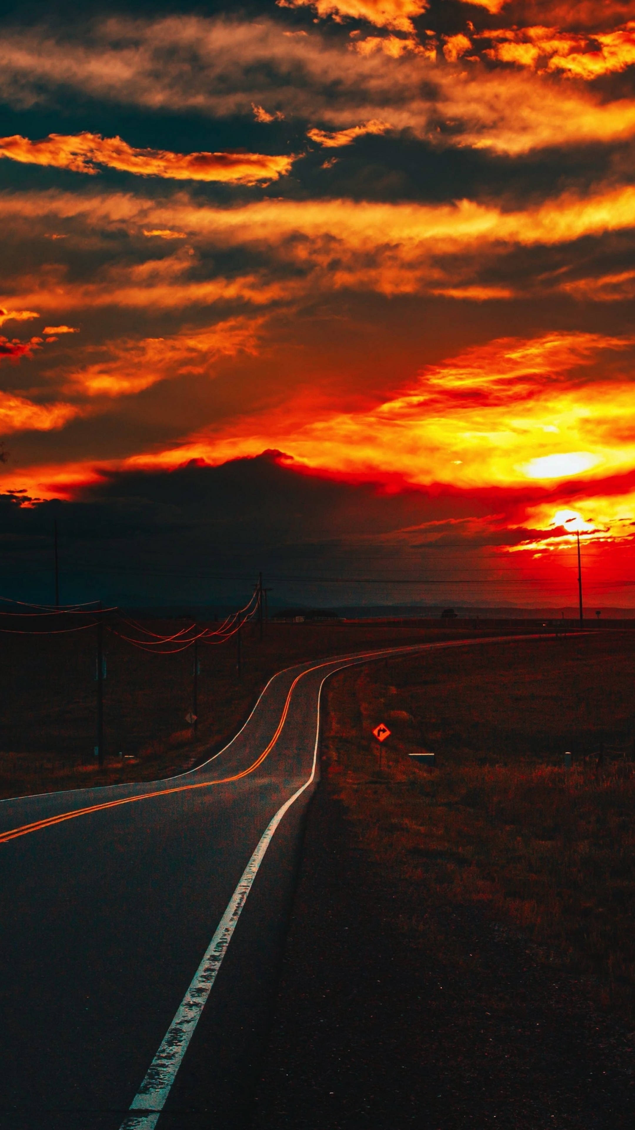 Sunset iPhone 桌布 壁紙 — 一條蜿蜒的道路穿過廣闊的田野，上方是帶有戲劇性雲彩的火紅橙色和紅色日落天空。