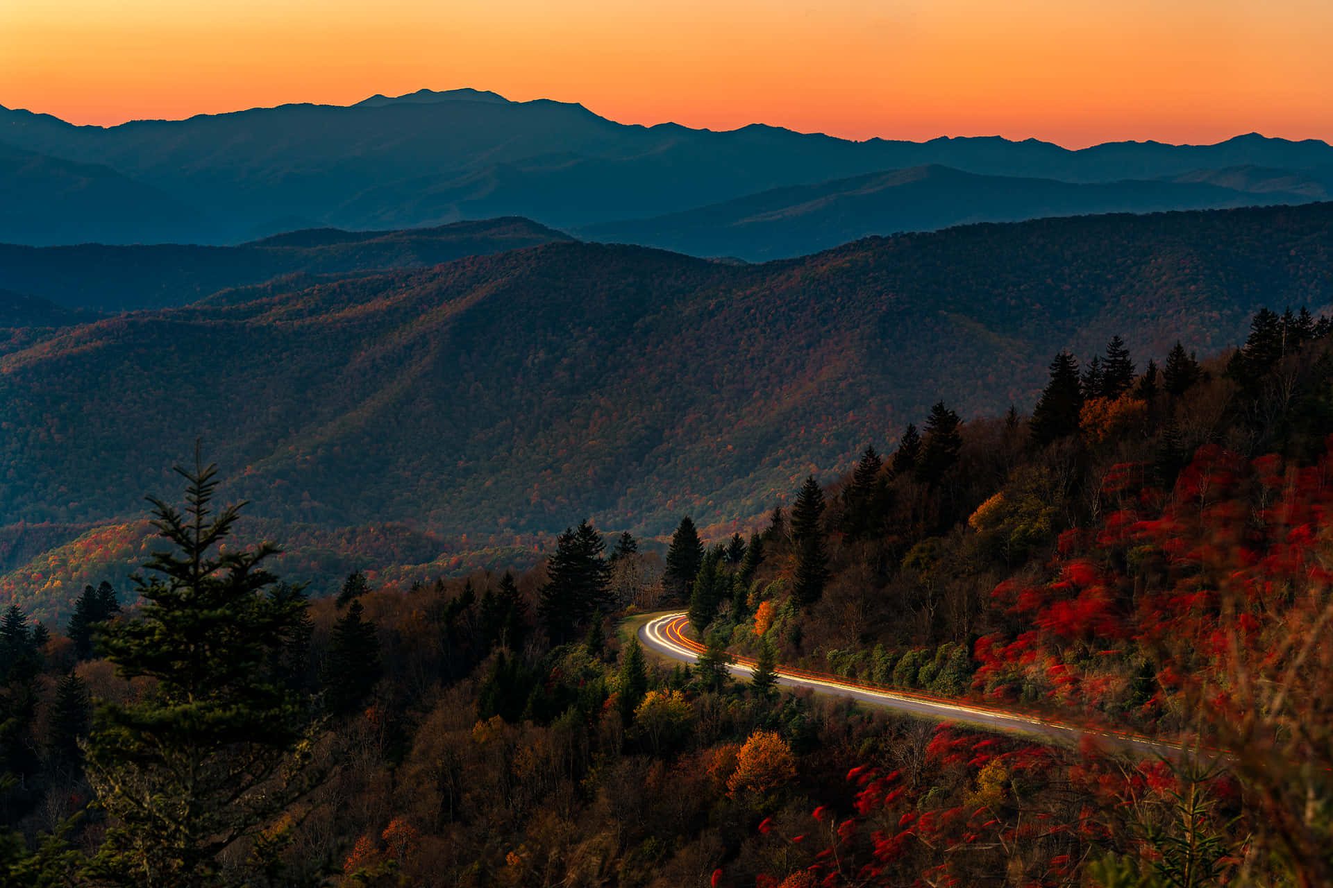 Blue Ridge Parkway 桌布 壁紙 — 一條蜿蜒的道路穿過秋日森林，上方是層層山脈之上火紅橙色的日落天空。