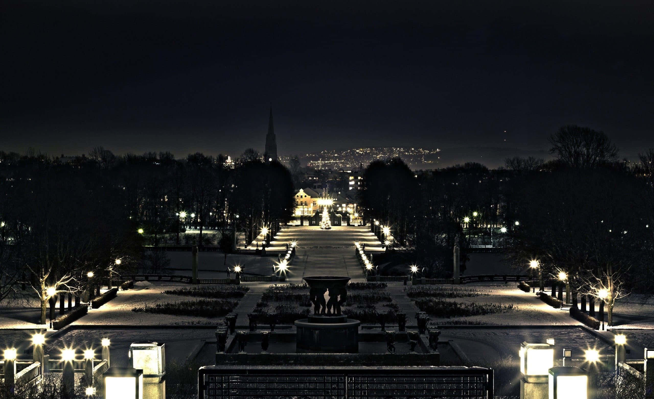 Winter Cityscape 桌布 壁紙 — 一條被雪覆蓋的公園小徑在夜晚被發光的燈籠照亮，通往黑暗天空下遠處的城市景觀。