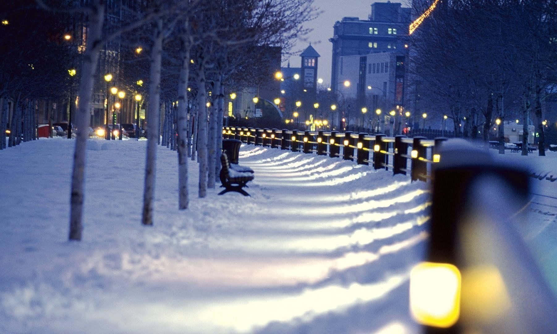 Winter Cityscape 桌布 壁紙 — 一條被雪覆蓋的城市公園小徑，夜晚被溫暖的路燈照亮，在純白的地面上投下長長的影子。