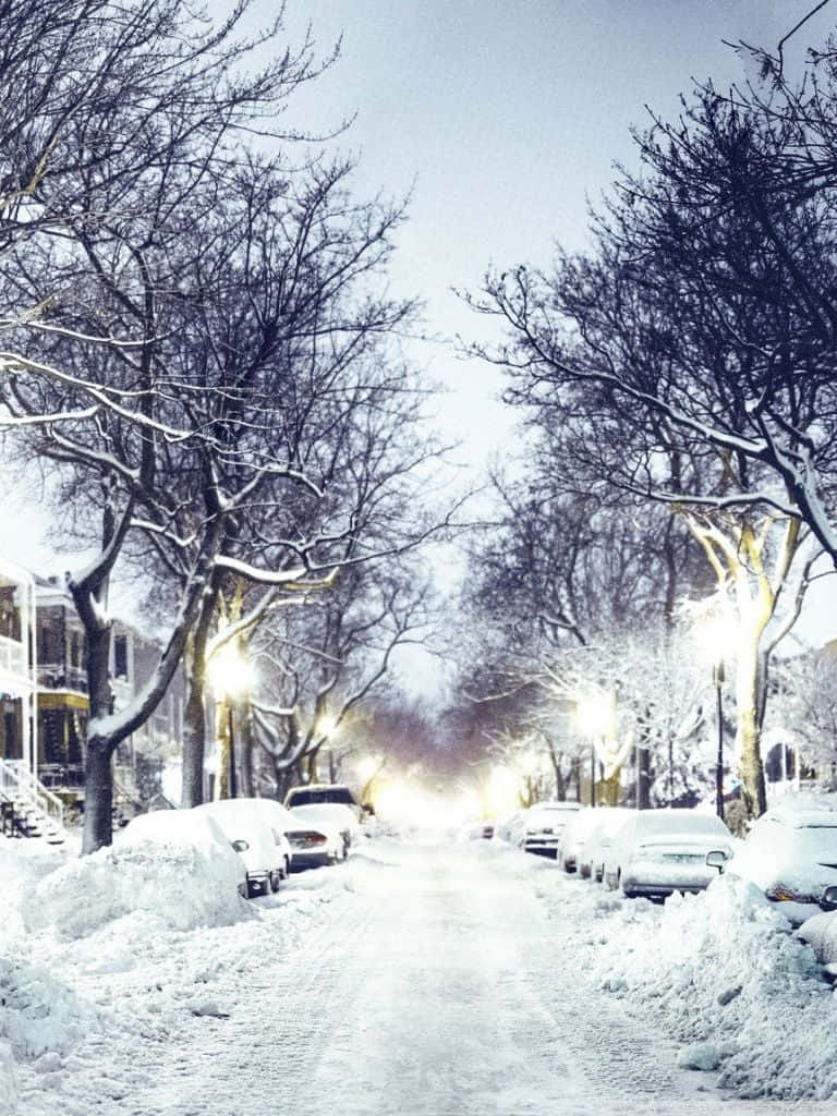 Winter Phone 桌布 壁紙 — 一條被雪覆蓋的城市街道，兩旁是光禿禿的樹木和停放的汽車，在暮色中閃爍的路燈下。