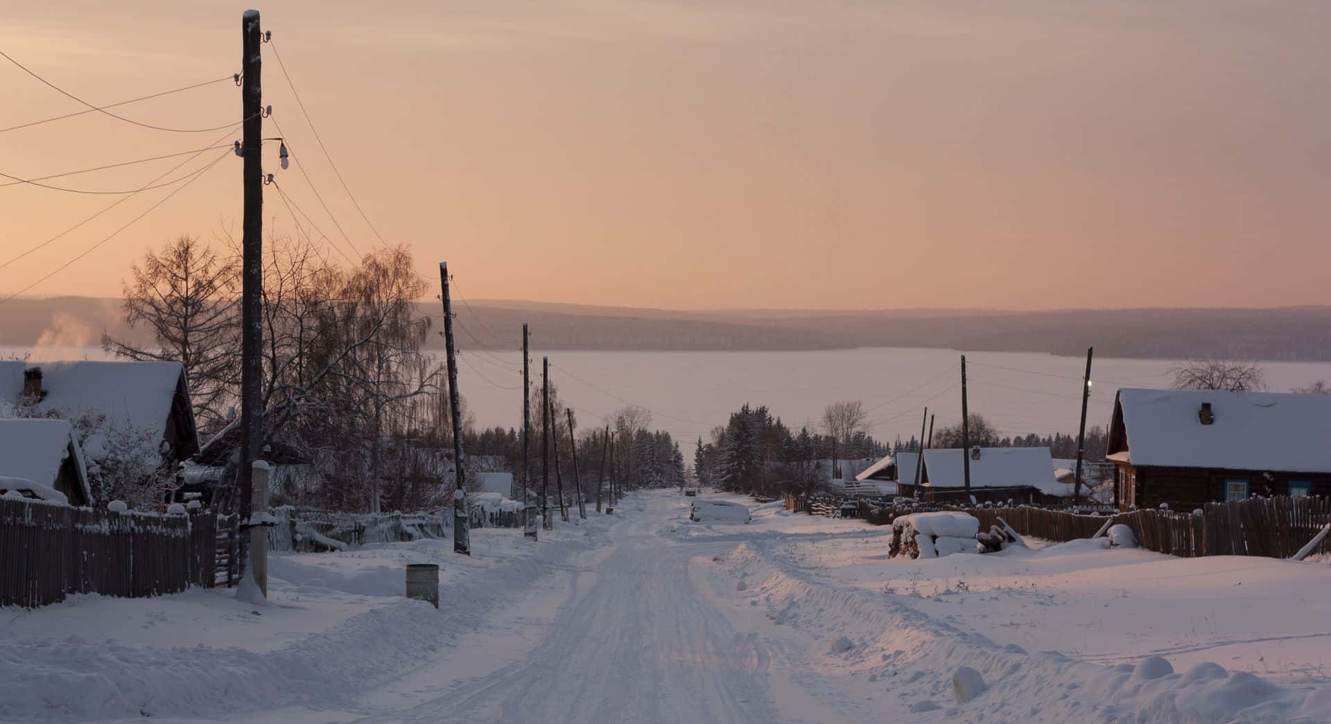 Snowy Village 桌布 壁紙 — 一條被雪覆蓋的村莊道路，在溫暖橙色天空和光禿樹木的映襯下，延伸向夕陽下的冰凍湖泊。
