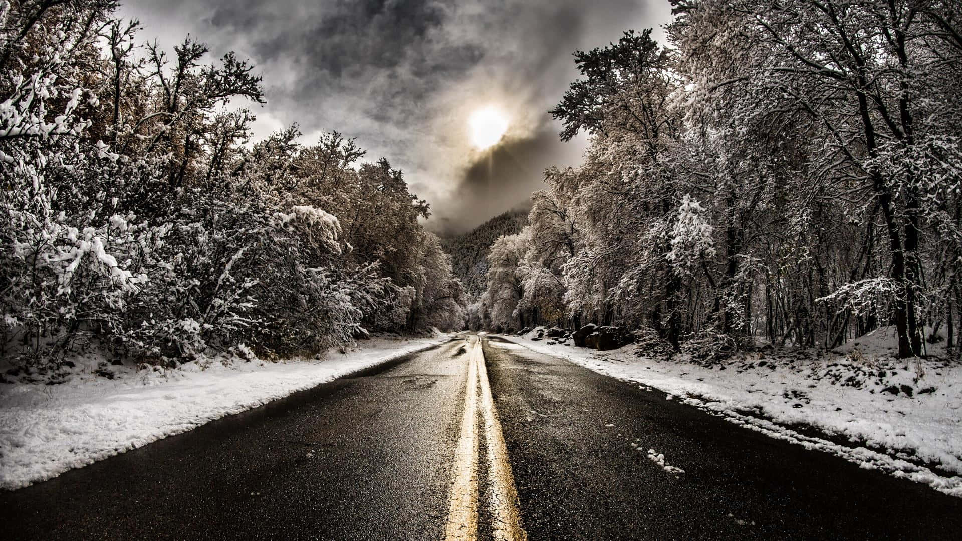 道路 桌布 壁紙 — 一條被雪覆蓋的道路蜿蜒穿過霜凍的森林，天空戲劇性地多雲，明亮的陽光穿透雲層。
