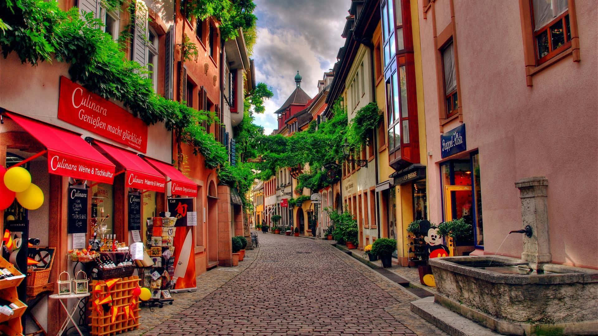 Germany 桌布 壁紙 — 一條迷人的鵝卵石街道，兩旁是色彩繽紛的建築和綠植，天空多雲。