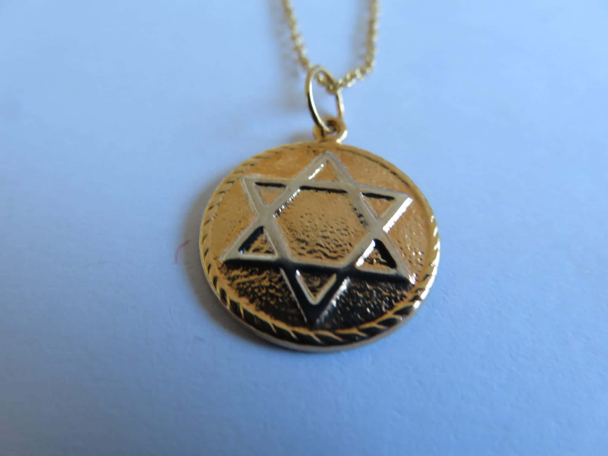 Thelema 桌布 壁紙 — 一條鏈上的金色大衛之星吊墜，靠在帶有細微紋理的柔和藍色背景上。