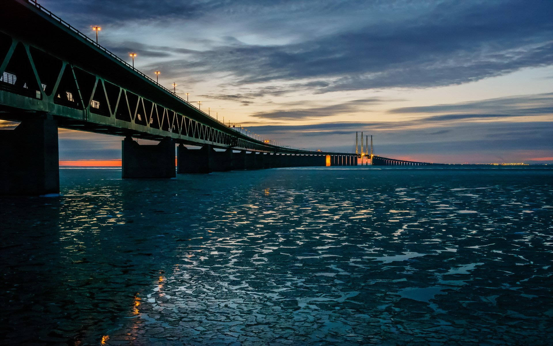 Sweden 桌布 壁紙 — 一條長橋橫跨冰冷的水面，在黃昏時分，溫暖的燈光反射在冰凍的表面上。