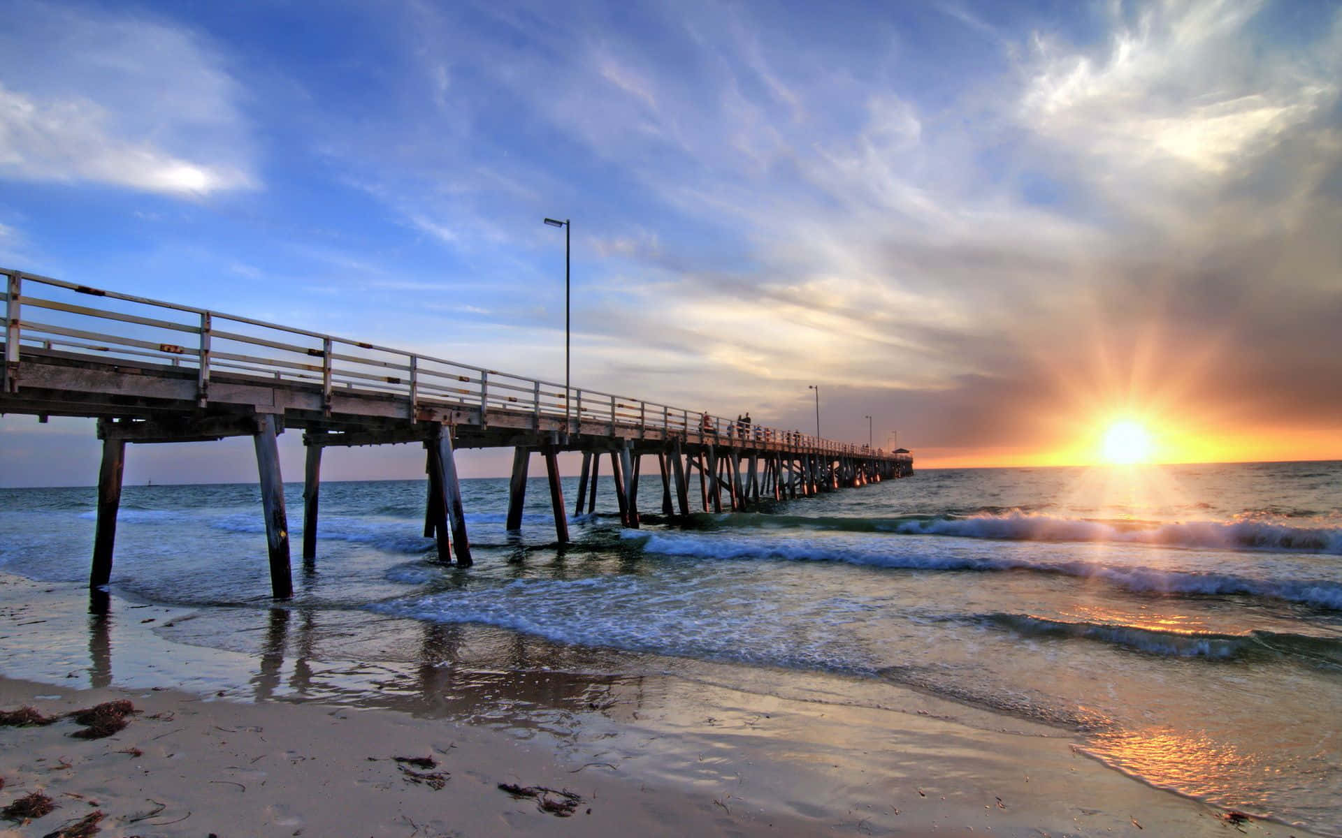 Australia Beach 桌布 壁紙 — 一條長長的木製碼頭在日落時延伸入海洋，金色的光線反射在波浪和濕沙上。