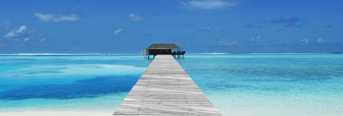 Maldives Island 桌布 壁紙 — 一條長長的木製碼頭延伸至清澈碧綠的海水中，上方是明亮的藍天，點綴著幾朵白雲。