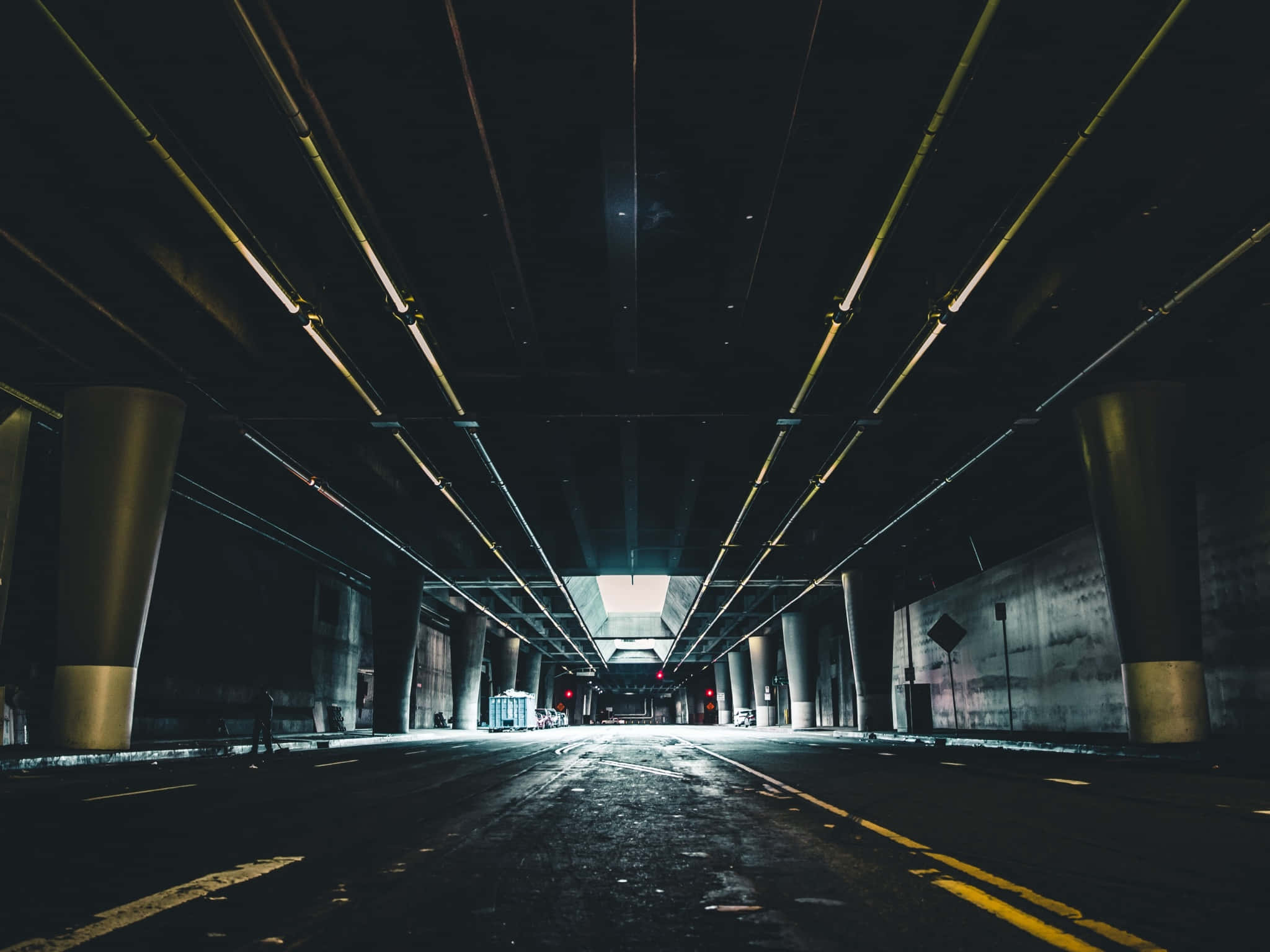 Dark Underground 桌布 壁紙 — 一條長長的黑暗隧道，有黃色管道和柱子，延伸至明亮的出口，可見汽車和紅燈。