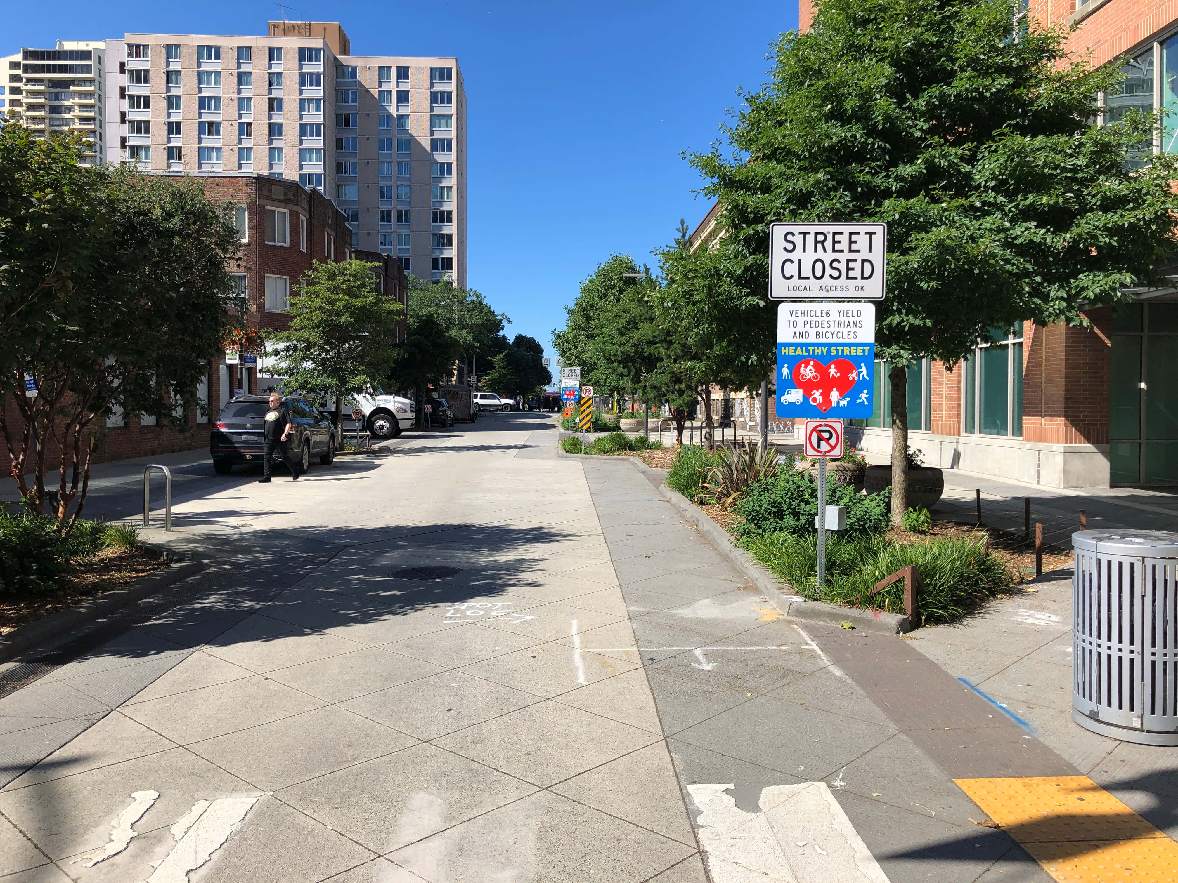 Street 桌布 壁紙 — 一條陽光普照的城市街道，禁止車輛通行，設有行人標誌，人行道兩旁種滿茂密的綠樹。