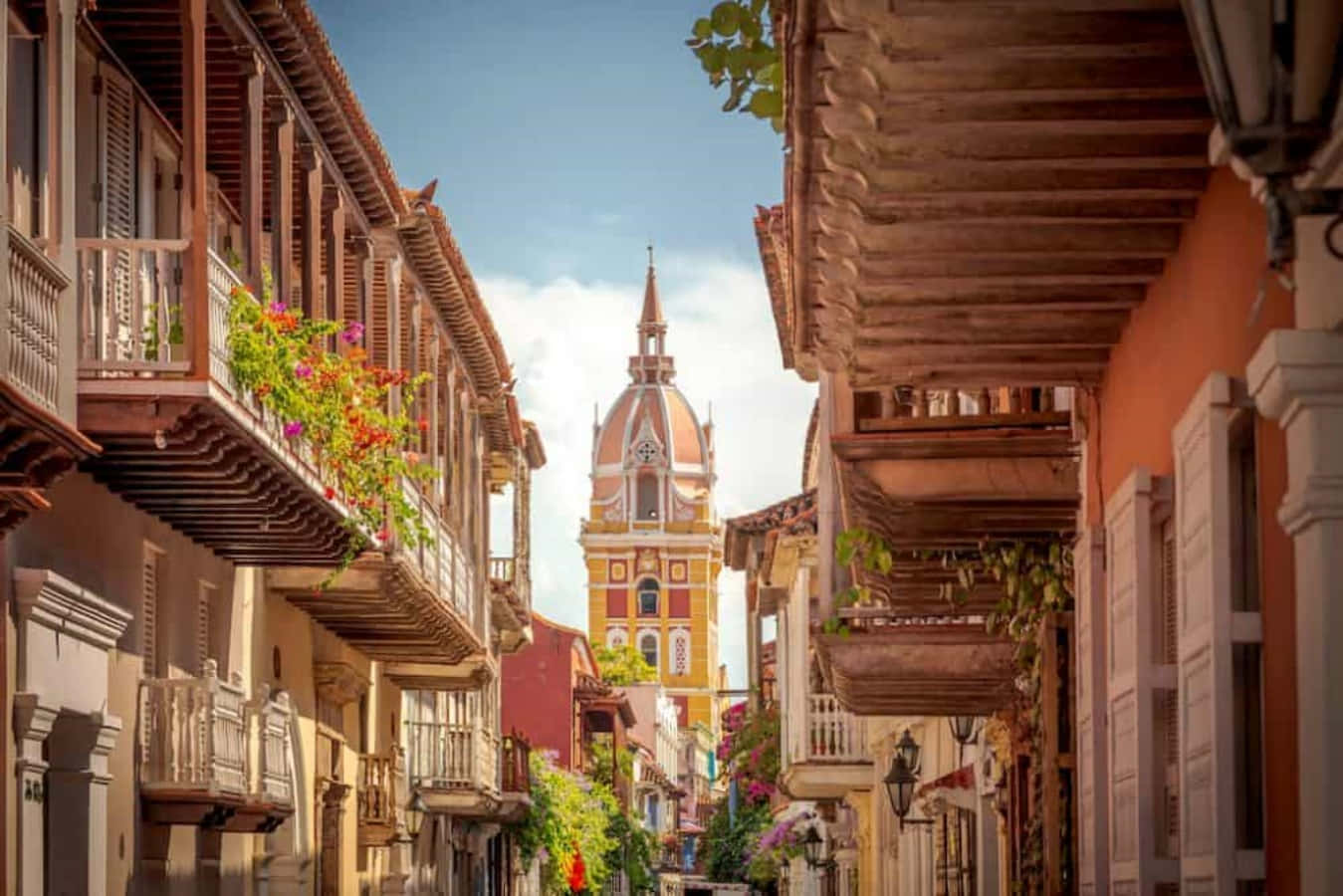 Colombia 桌布 壁紙 — 一條陽光普照的殖民風格街道，兩旁是木製陽台，陽台上開滿了色彩繽紛的花朵，通往一座充滿活力的教堂塔樓。