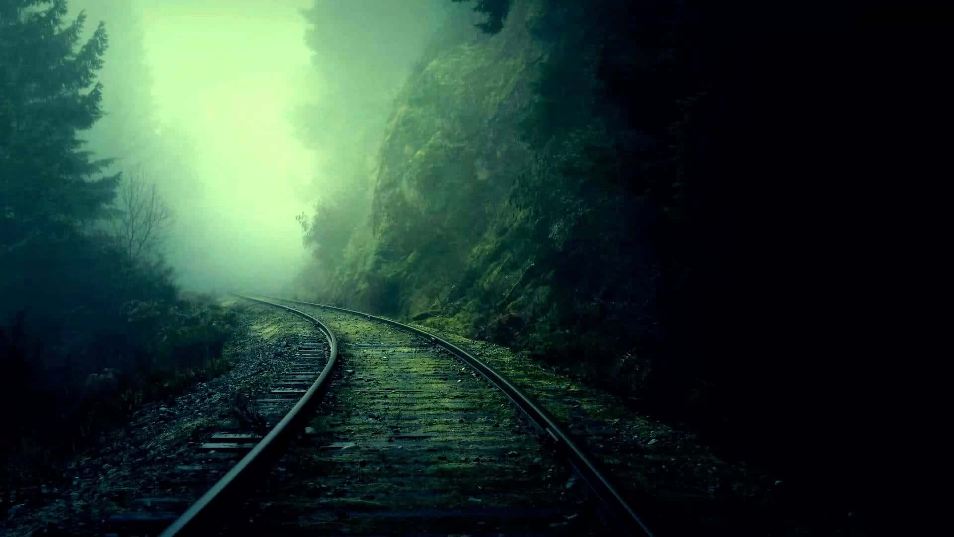Ambience 桌布 壁紙 — 一條霧氣瀰漫的鐵路軌道彎曲進入一條發光的綠色森林隧道，兩旁是黑暗的樹木和長滿苔蘚的岩石。