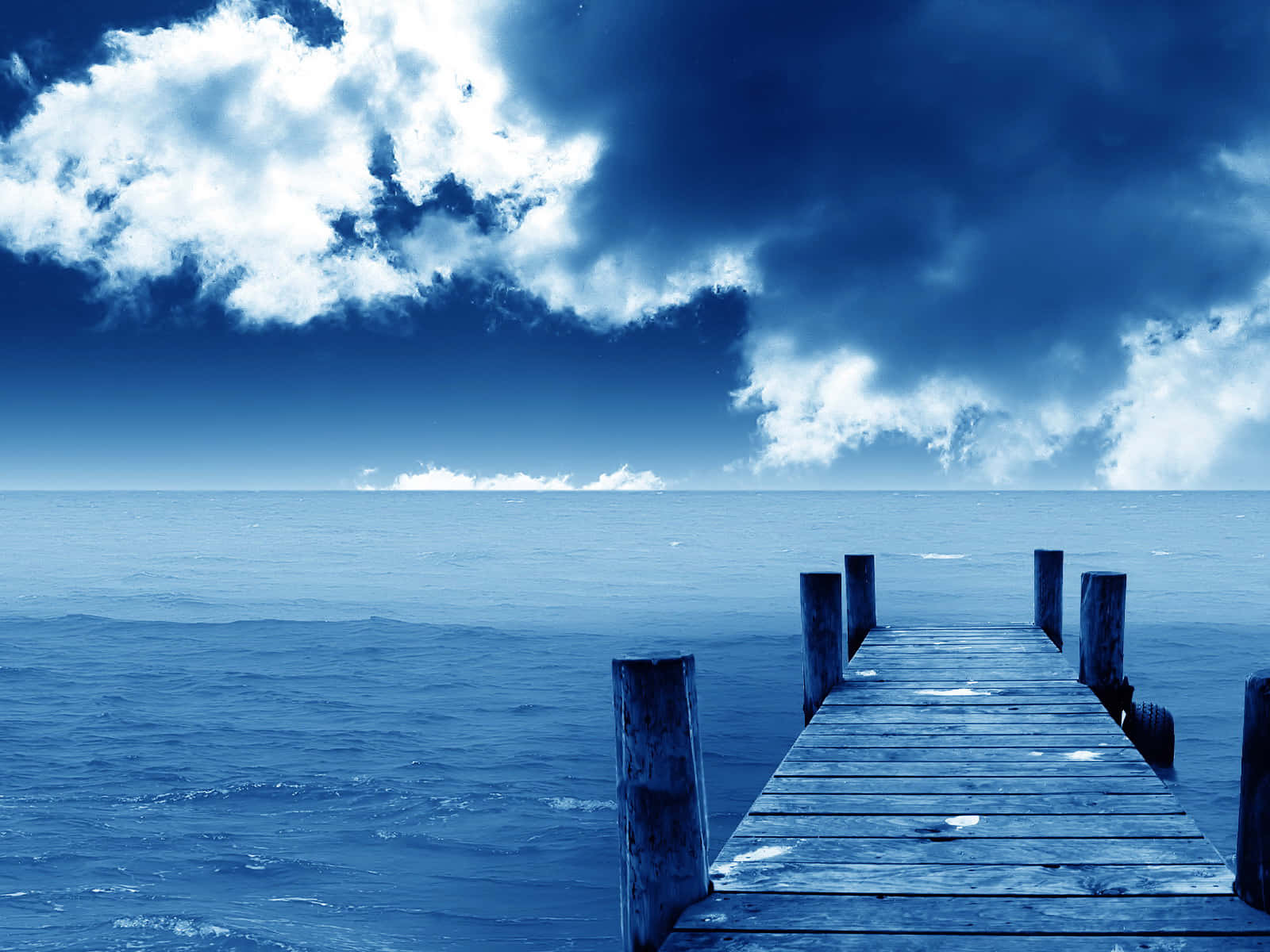 Blue Skies 桌布 壁紙 — 一條風化的木碼頭在戲劇性的多雲天空下延伸至廣闊的藍色大海。