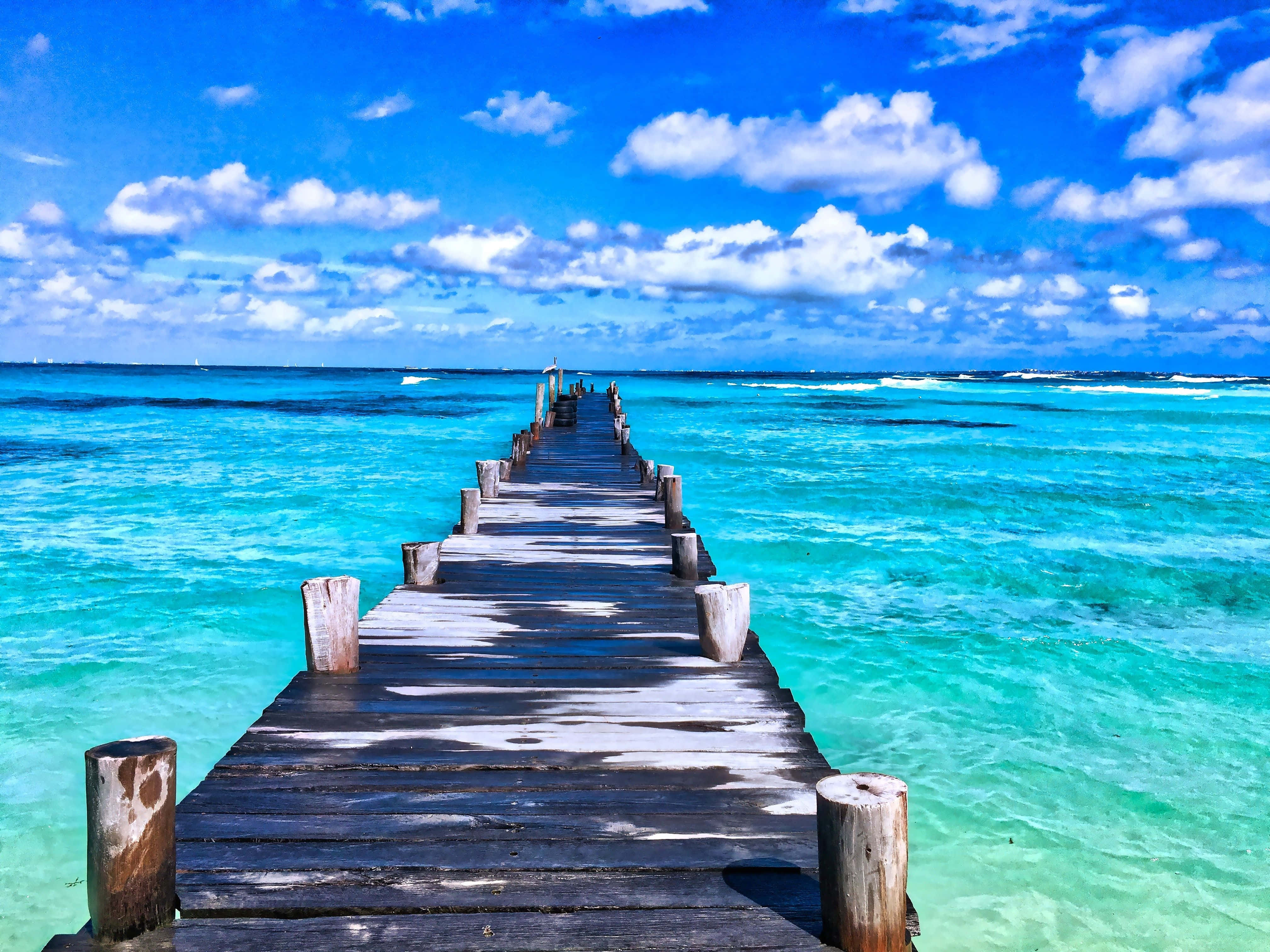 Beautiful Beach 桌布 壁紙 — 一條飽經風霜的木製碼頭延伸至充滿活力的碧綠海水，上方是點綴著蓬鬆白雲的明亮藍天。