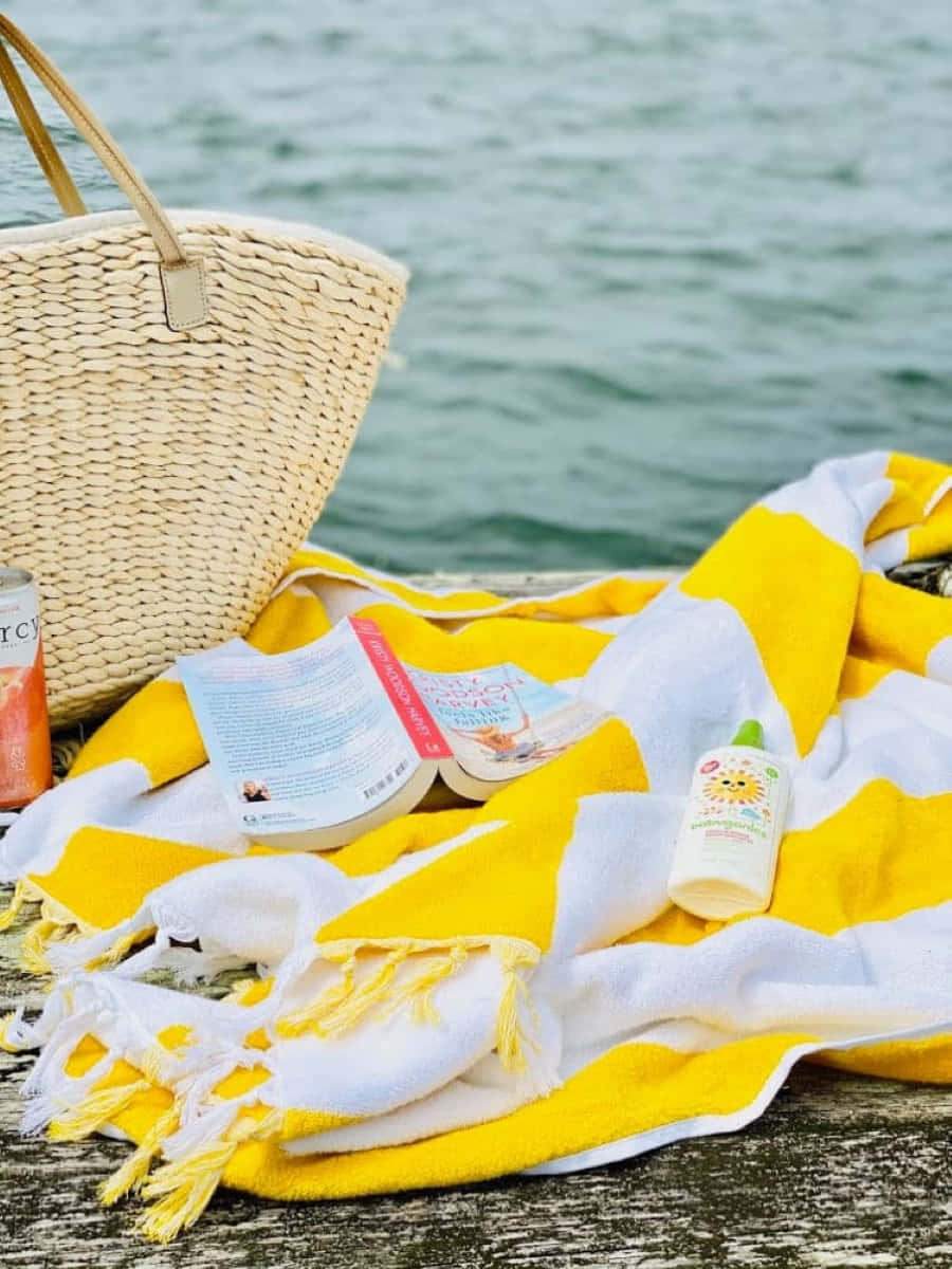 Beach Accessories 桌布 壁紙 — 一條黃白條紋的沙灘毛巾帶有流蘇，放在木製碼頭上，旁邊有一個編織籃、書本、防曬霜和一罐飲料，背景有水。