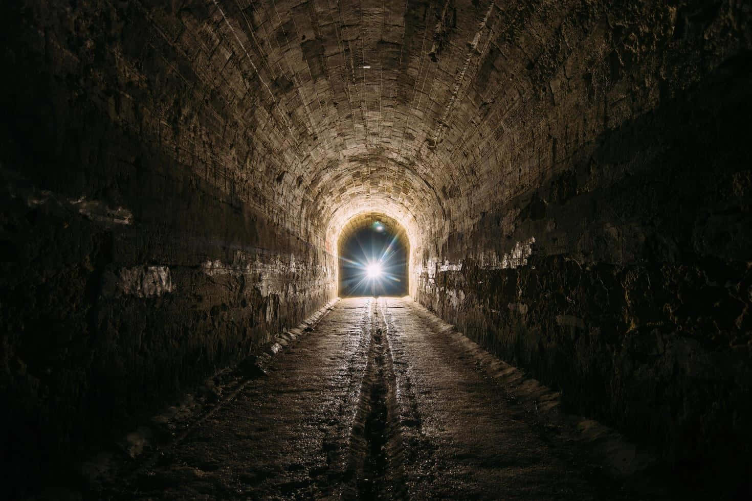 Dark Underground 桌布 壁紙 — 一條黑暗的石頭隧道，地面潮濕，通往遠端明亮的發光出口。