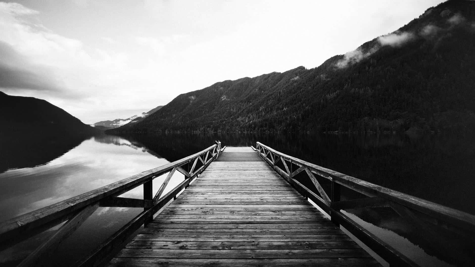 Black And White Nature 桌布 壁紙 — 一條黑白相間的木製碼頭伸向一個平靜的湖泊，四周被霧氣籠罩的森林山脈環繞，天空多雲。