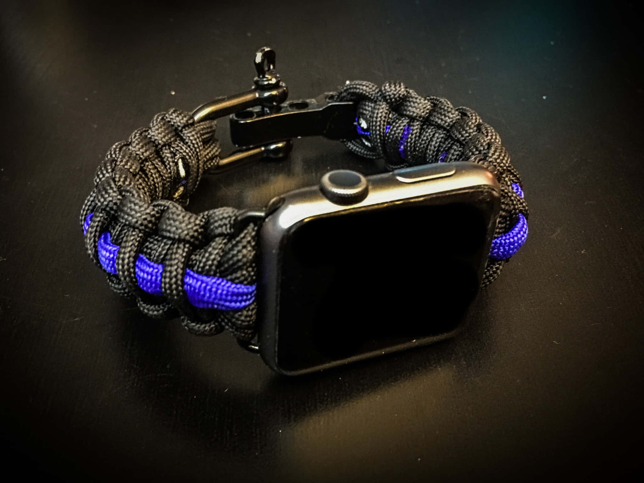 細藍線 桌布 壁紙 — 一條黑色和藍色的傘繩Apple Watch錶帶放在黑暗的表面上，在聚焦的燈光下。