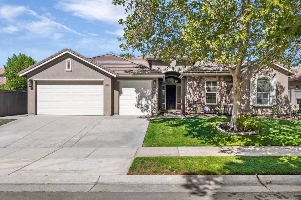 1561 Aimwell Avenue, Sacramento 壁紙 — 一棟單層郊區住宅，擁有石頭外牆和白色車庫門，矗立在明亮的藍天下，周圍是茂密的綠草，一棵大樹投下斑駁的陰影。