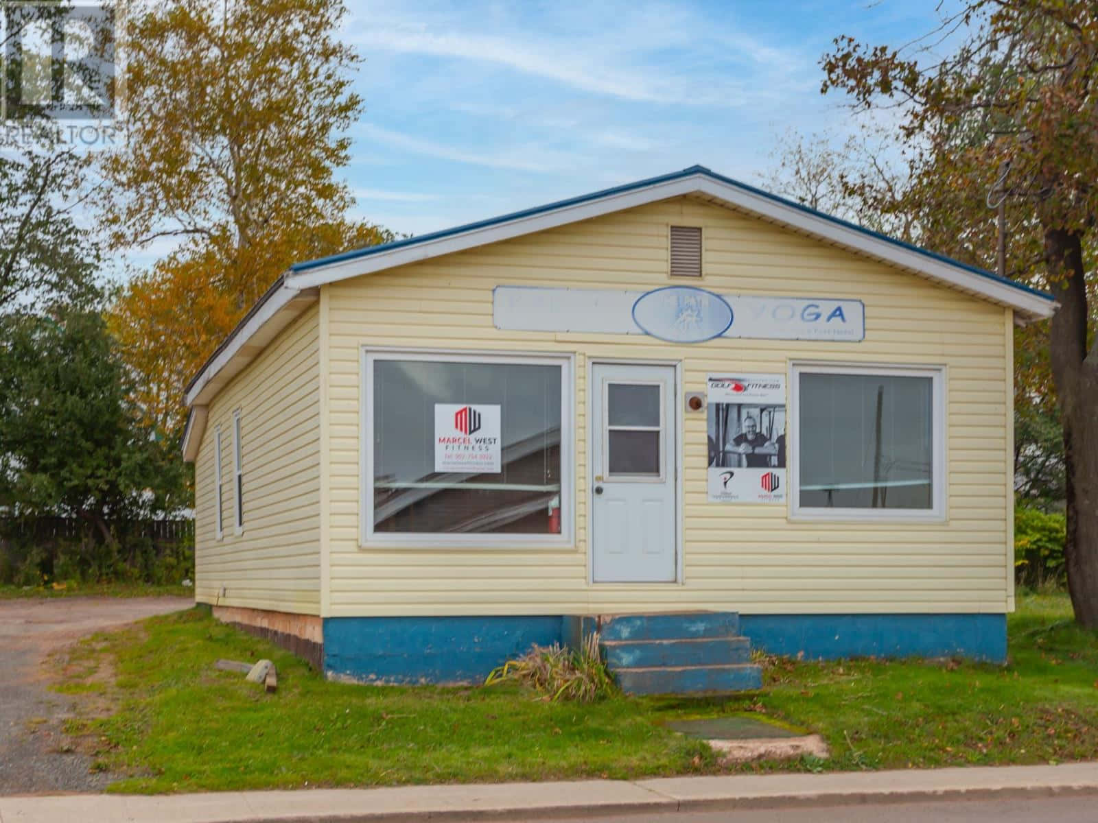 Charlottetown 桌布 壁紙 — 一棟淺黃色的單層建築，帶有藍色飾邊和瑜伽工作室標誌，矗立在部分多雲的天空下，周圍環繞著秋樹。