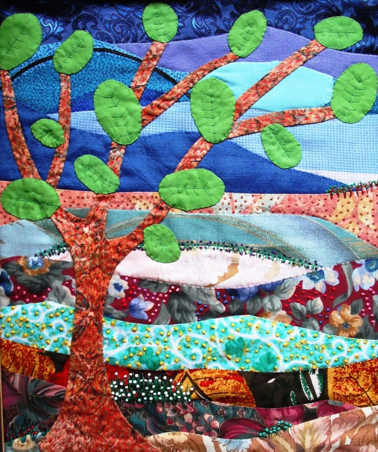 Textile Art 桌布 壁紙 — 一棵充滿活力的拼布樹，帶有綠色葉子貼花，矗立在藍色和紅色的拼布天空前，帶有縫線細節。