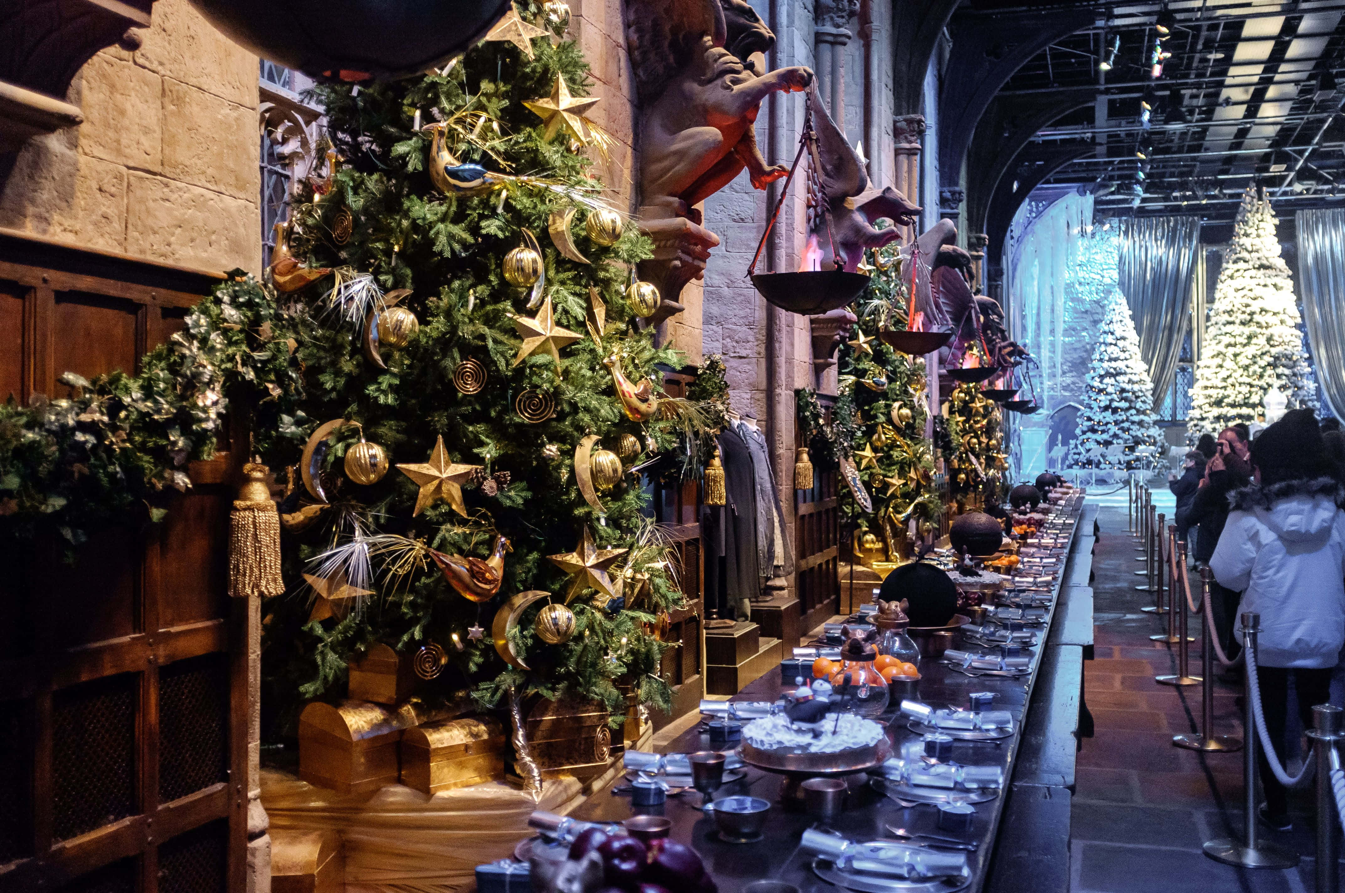 Great Hall Harry Potter Desktop 桌布 壁紙 — 一棵大聖誕樹裝飾著金色星星和飾品，矗立在石廳的長餐桌旁，有訪客在觀看。