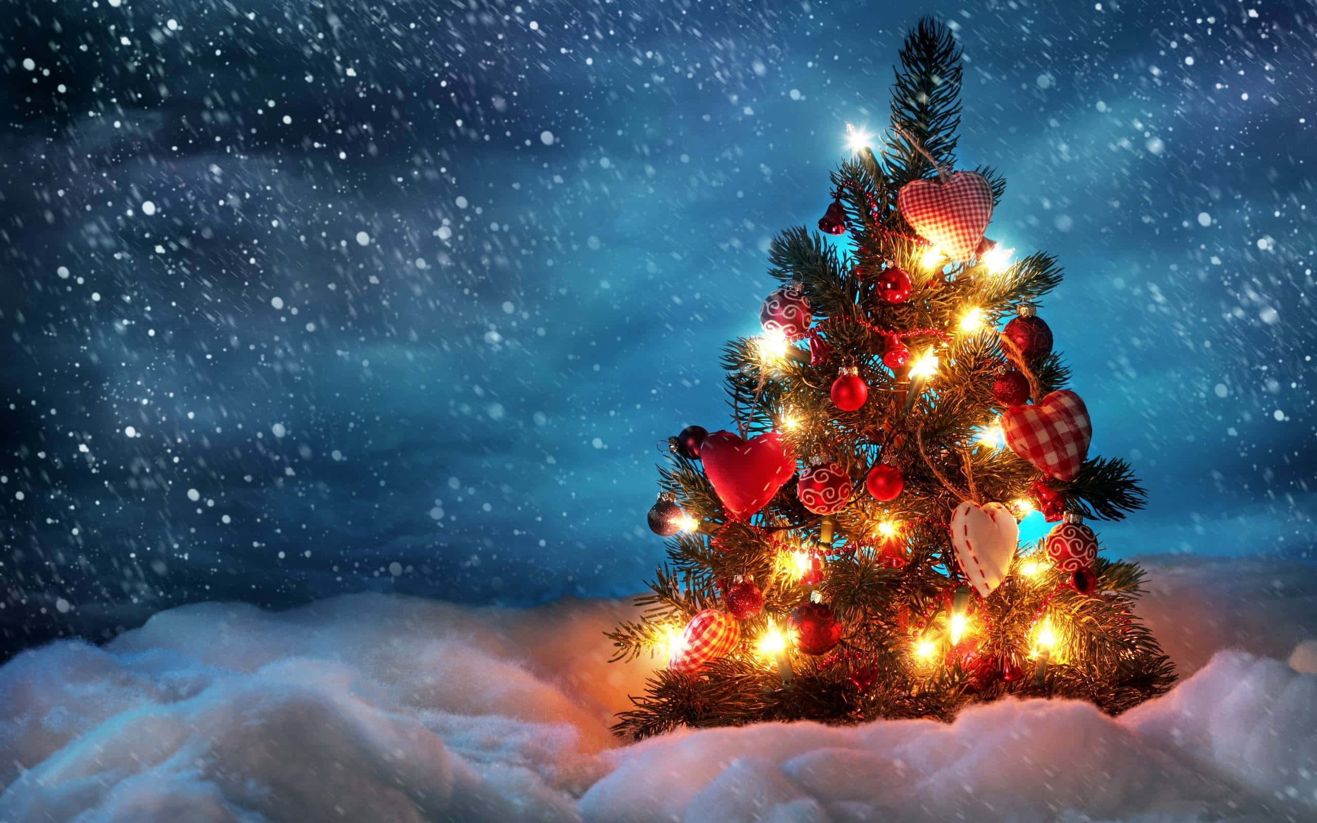 Snowy Christmas 桌布 壁紙 — 一棵溫暖照亮的聖誕樹，裝飾著紅色心形和飾品，矗立在雪地中，背景是飄雪的夜空。
