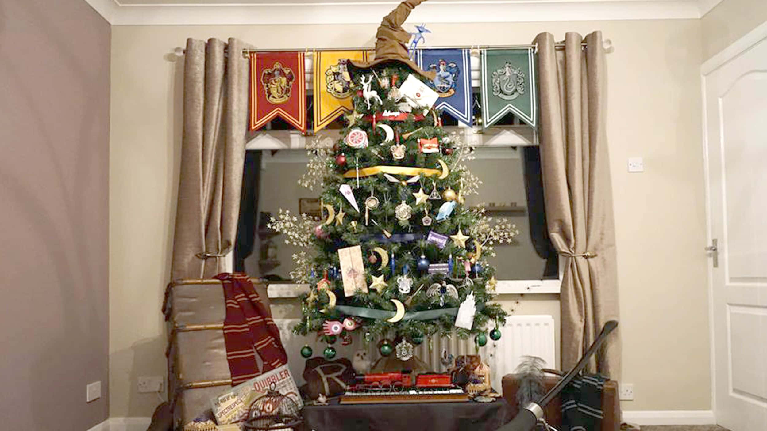 Harry Potter Christmas 桌布 壁紙 — 一棵裝飾著哈利波特主題飾品和橫幅的聖誕樹掛在一個有窗戶和窗簾的舒適客廳裡。
