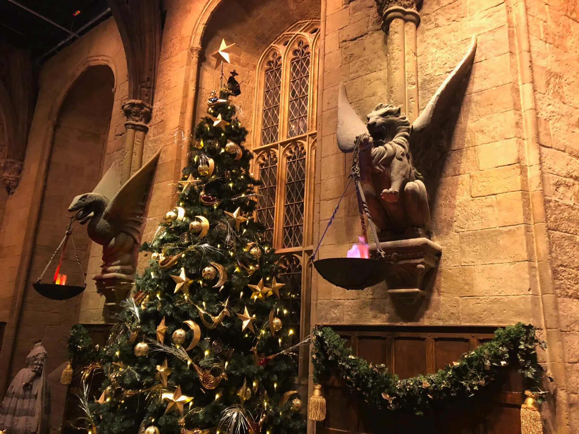 Harry Potter Christmas 桌布 壁紙 — 一棵裝飾著金色星星和飾品的大聖誕樹矗立在石造大教堂中，教堂內有手持發光燈籠的石像鬼雕像。